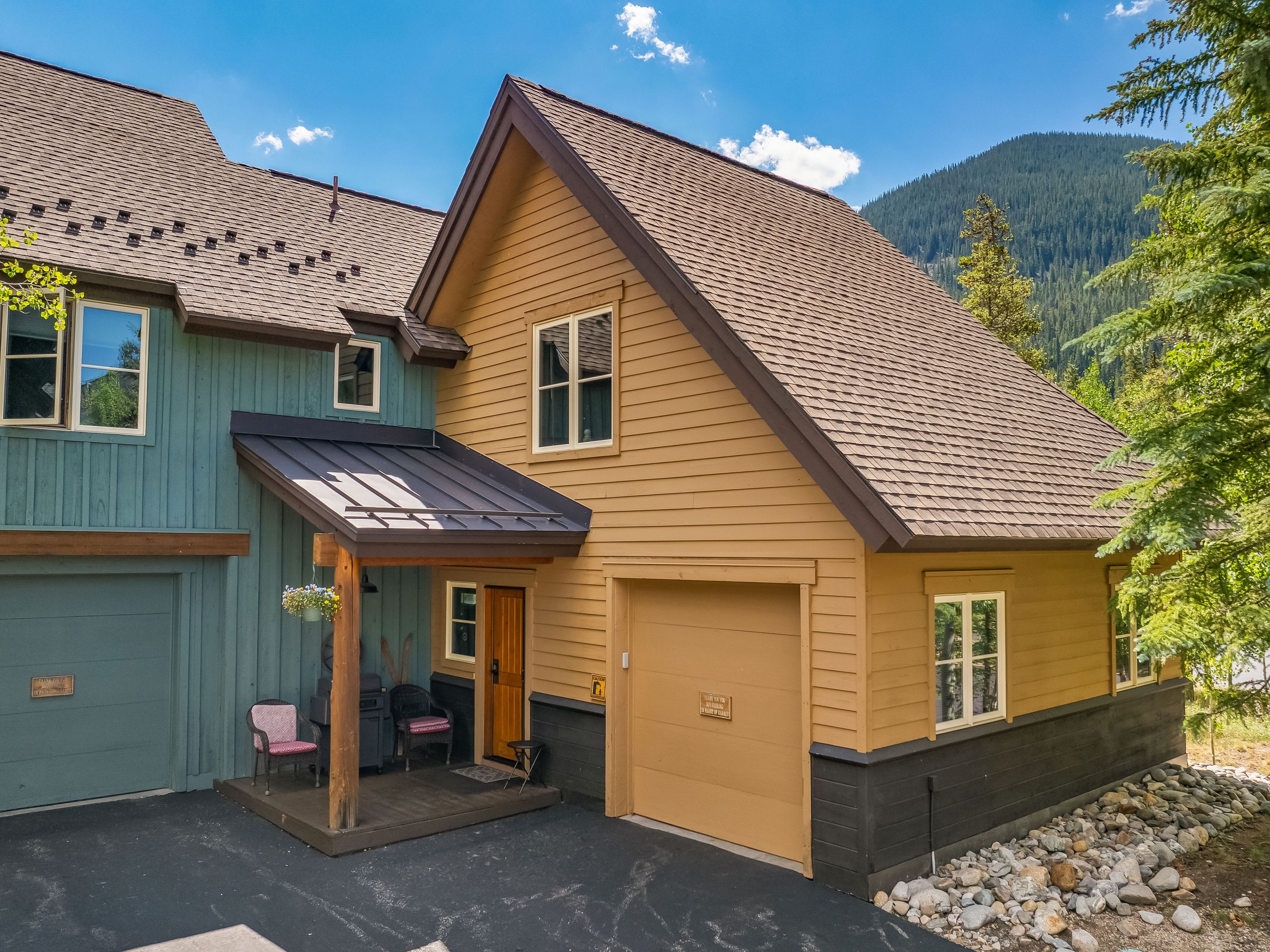 This exterior view captures a multi-level mountain home featuring a blend of teal and tan horizontal siding with dark trim accents. The property includes a covered entryway with a wooden porch, a single-car garage, and a rocky landscape border, all set against a backdrop of lush pine trees and a clear blue sky. The architectural style is rustic and inviting, emphasizing a cozy, cabin-like aesthetic suitable for a mountain retreat.