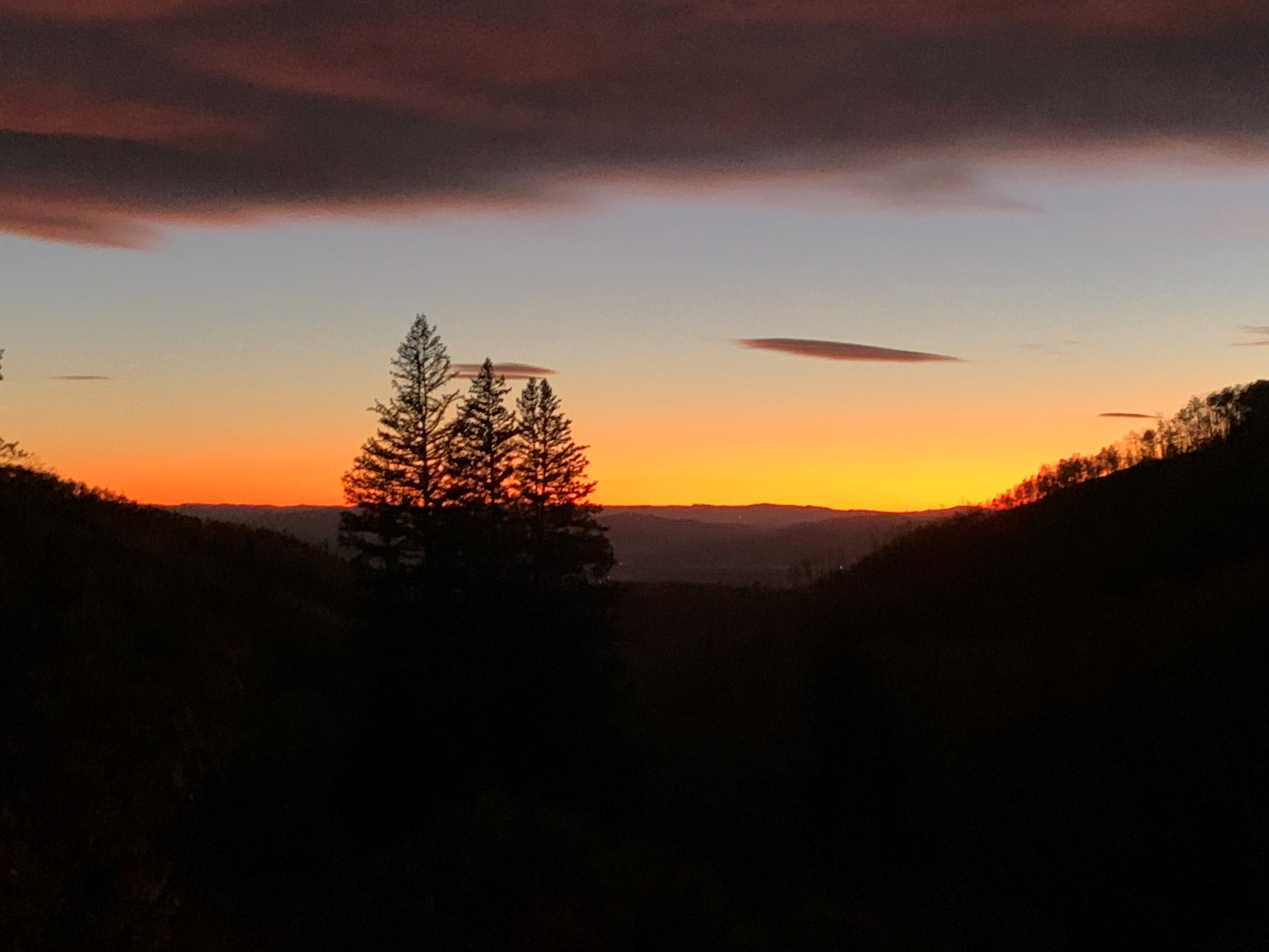 This aerial view captures a serene landscape at sunset, with silhouetted trees in the foreground and rolling hills fading into the horizon. The sky is painted with vibrant hues of orange and purple, creating a dramatic and picturesque scene. The image evokes a sense of tranquility and natural beauty, highlighting the potential for a peaceful and secluded property.