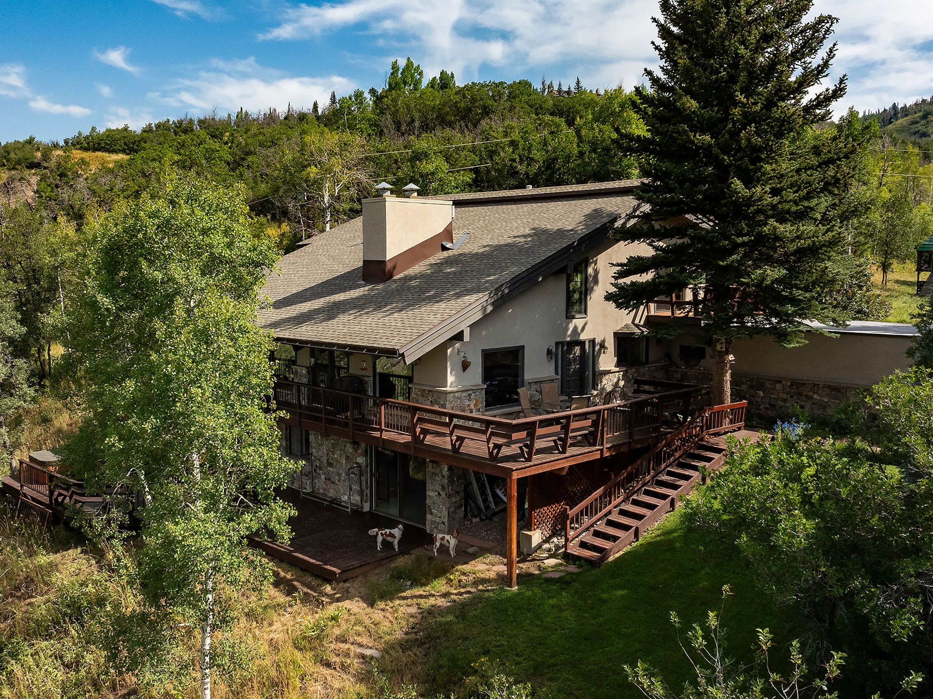 This aerial view showcases a beautiful multi-level home with a large wooden deck and stone accents. The house is nestled among lush greenery and mature trees, creating a sense of privacy and tranquility. The architecture blends seamlessly with the natural surroundings, offering a serene and inviting atmosphere.