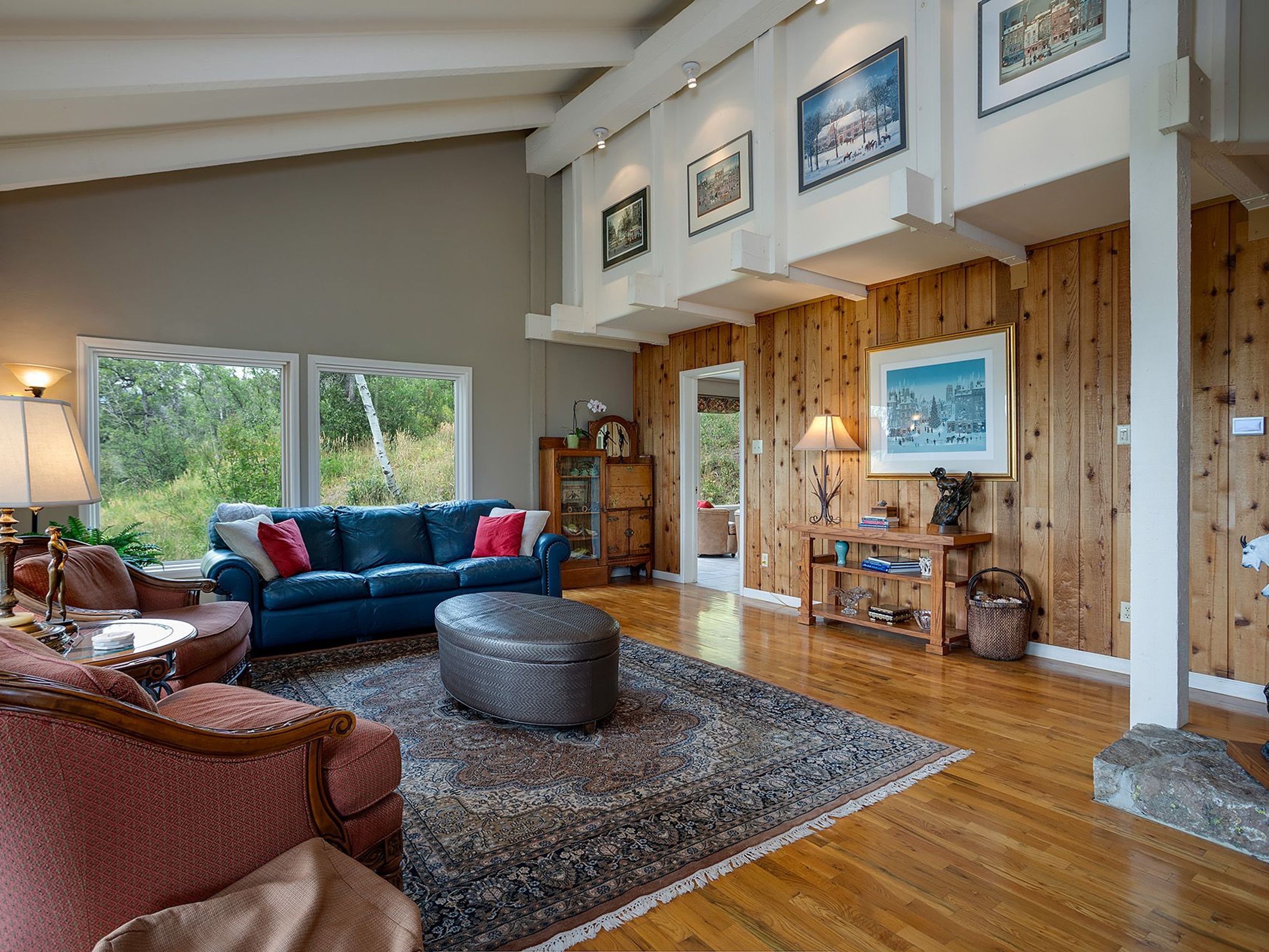 This is an interior shot of a living room featuring a blue leather sofa with red pillows, a large patterned area rug, and a brown leather ottoman. The room has wood-paneled walls, hardwood floors, and large windows that provide natural light and a view of the outdoors. The ceiling is high with exposed beams, and artwork is displayed on the walls, creating a warm and inviting atmosphere.