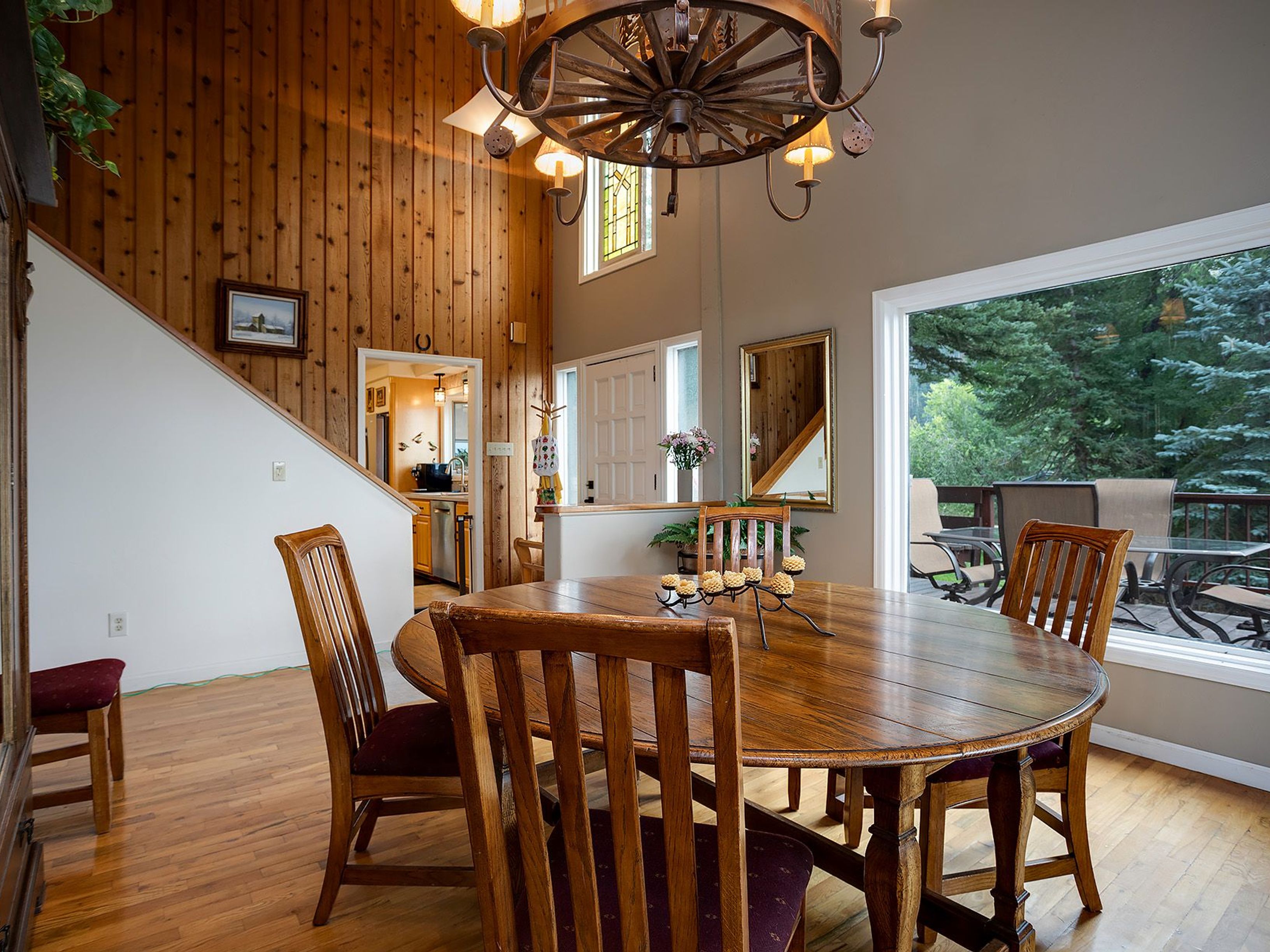 This is an inviting dining room featuring a large, round wooden table surrounded by wooden chairs with burgundy cushions. A unique wagon wheel chandelier hangs above the table, providing warm lighting. The room has wood-paneled walls and hardwood floors, creating a rustic and cozy atmosphere, with a large window offering a view of the outdoors.