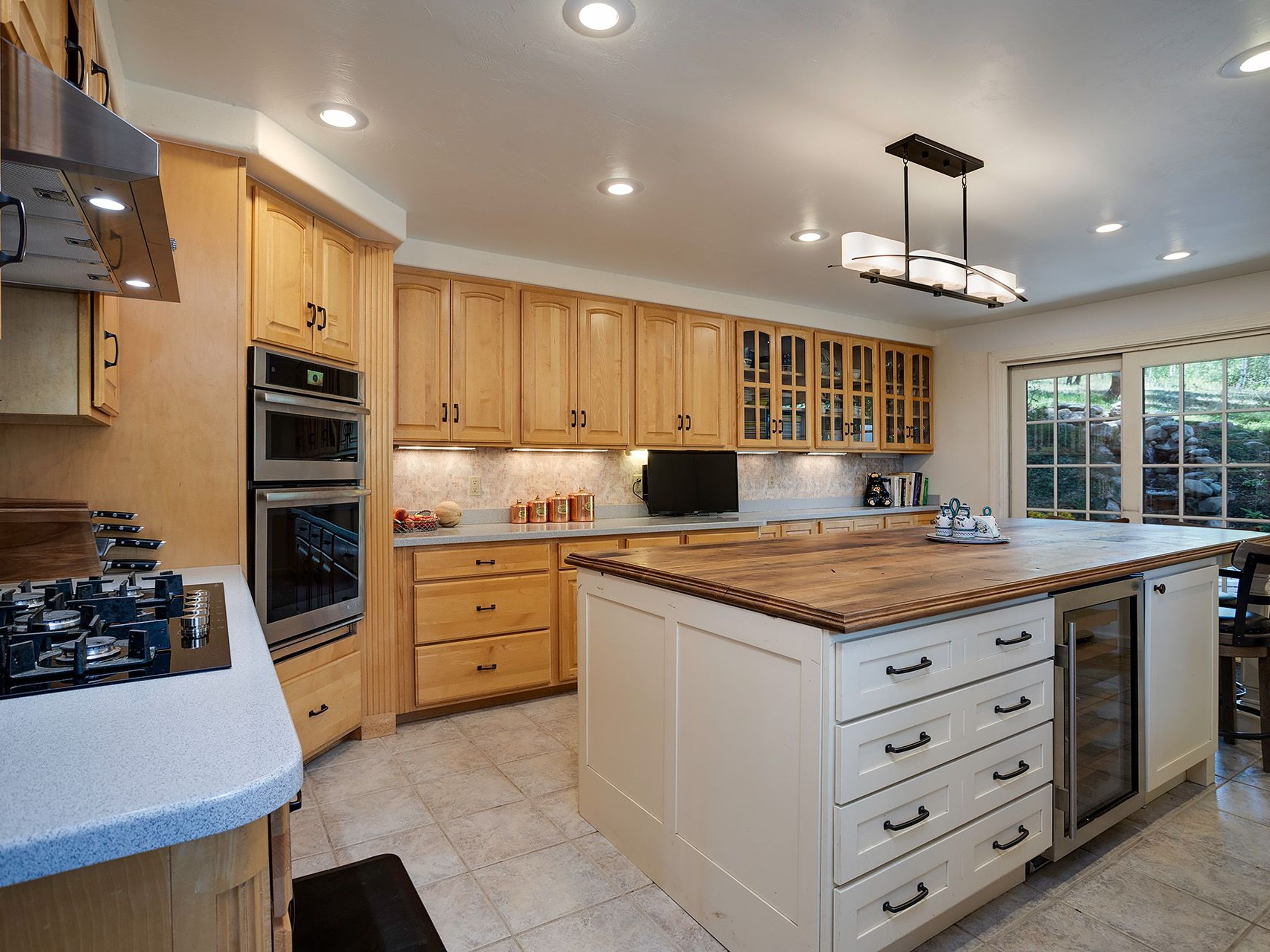 This is a well-lit kitchen featuring light wood cabinetry and a large center island with a butcher block countertop. Stainless steel appliances, including double ovens and a wine cooler, add modern convenience. The kitchen has a warm and inviting atmosphere, enhanced by recessed lighting and a view to the outside.