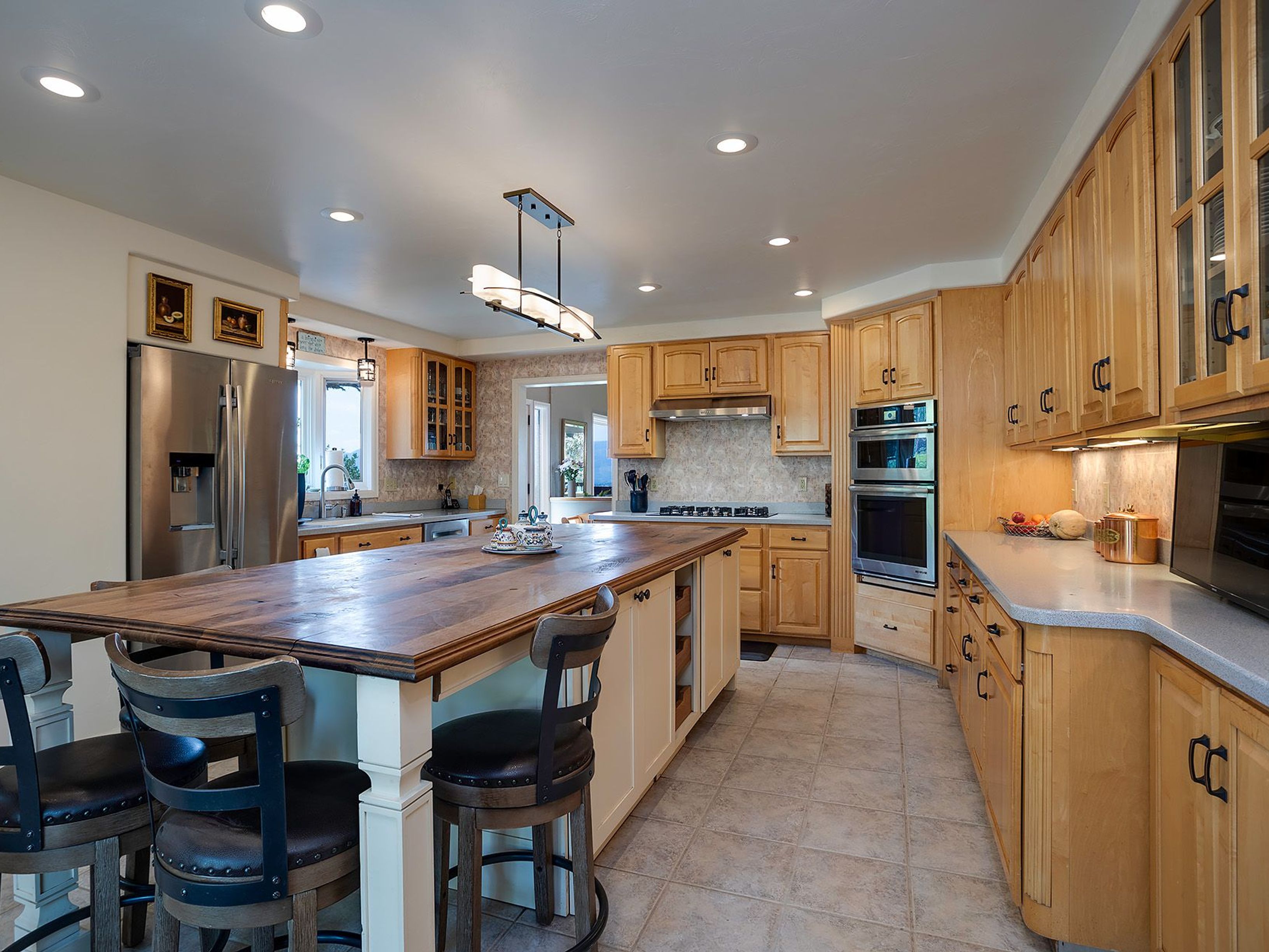 This is a spacious kitchen featuring light wood cabinetry, stainless steel appliances, and a large center island with a wooden countertop and seating. The kitchen has a warm and inviting feel, with tile flooring and recessed lighting throughout. The layout provides ample counter space and storage, making it ideal for cooking and entertaining.