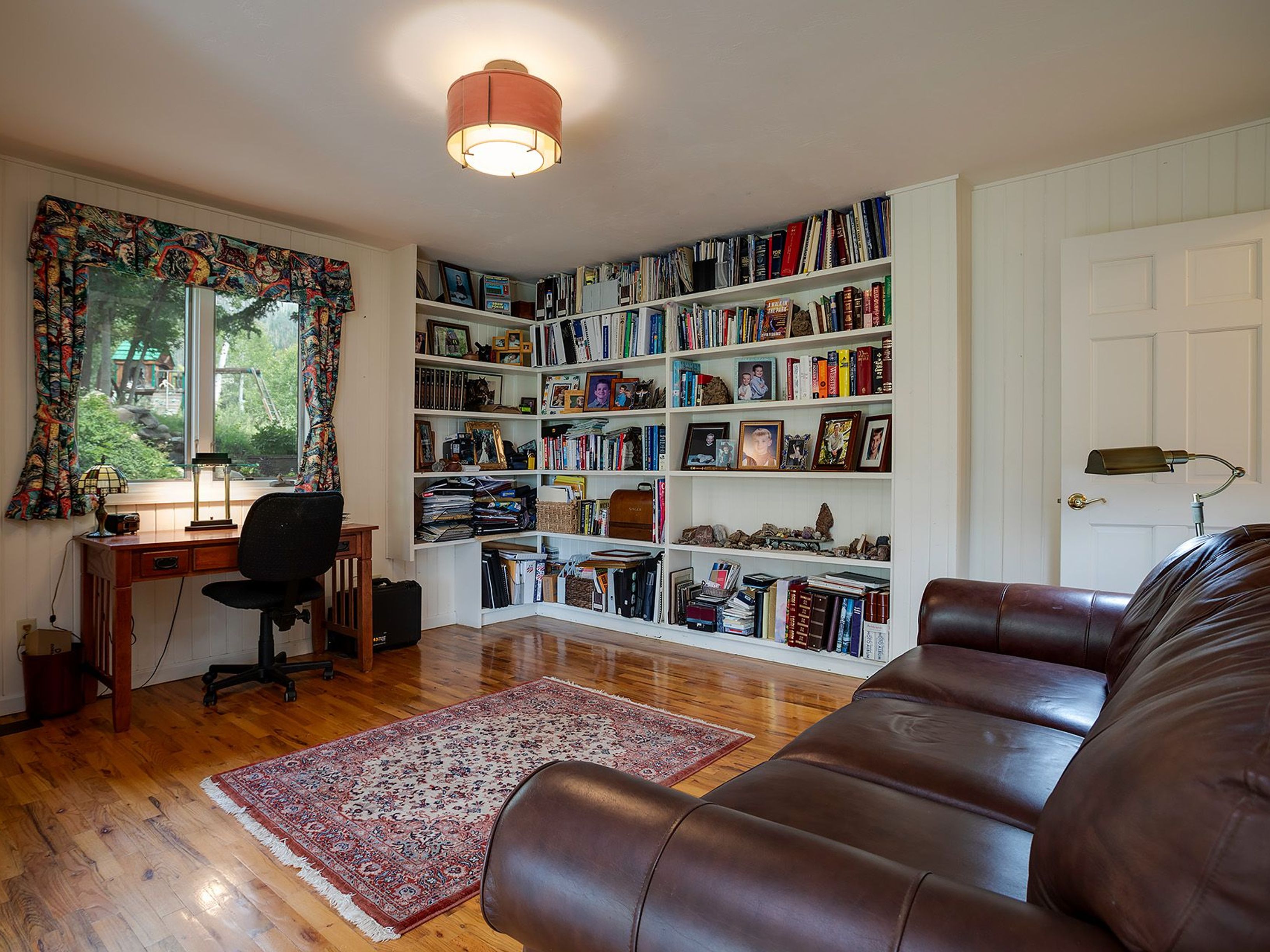 This is an interior shot of a home office or study featuring a large built-in bookshelf filled with books and decorative items. A wooden desk with a chair sits near a window with patterned curtains, and a brown leather couch is positioned in the foreground. The room has hardwood floors and a warm, inviting atmosphere.