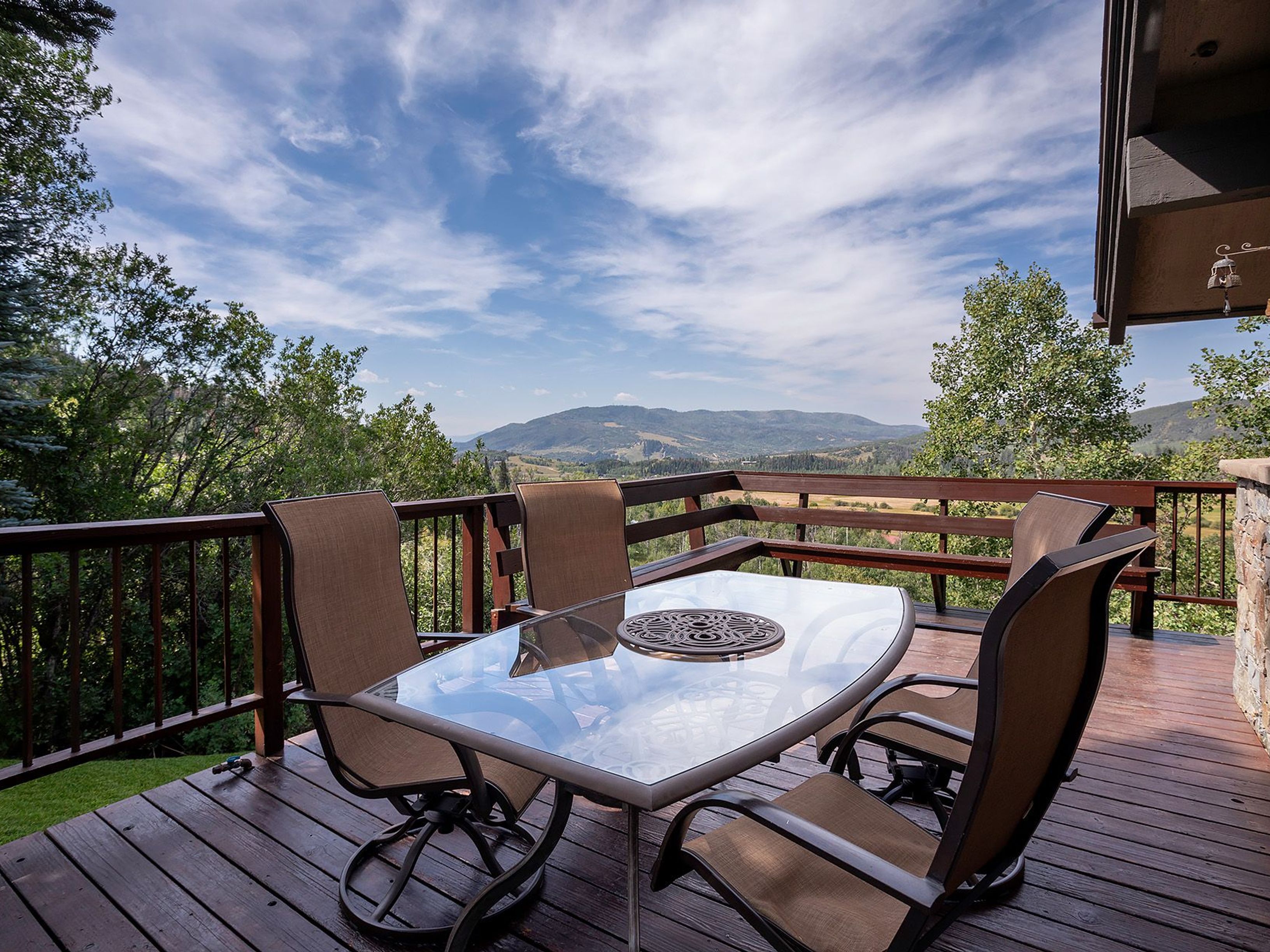 This image showcases a beautifully arranged outdoor dining area on a wooden deck, offering a stunning view of rolling hills and a partly cloudy sky. The glass-topped table and comfortable chairs create an inviting space for relaxation and entertainment. The wooden railing adds a touch of rustic charm, blending seamlessly with the natural surroundings.