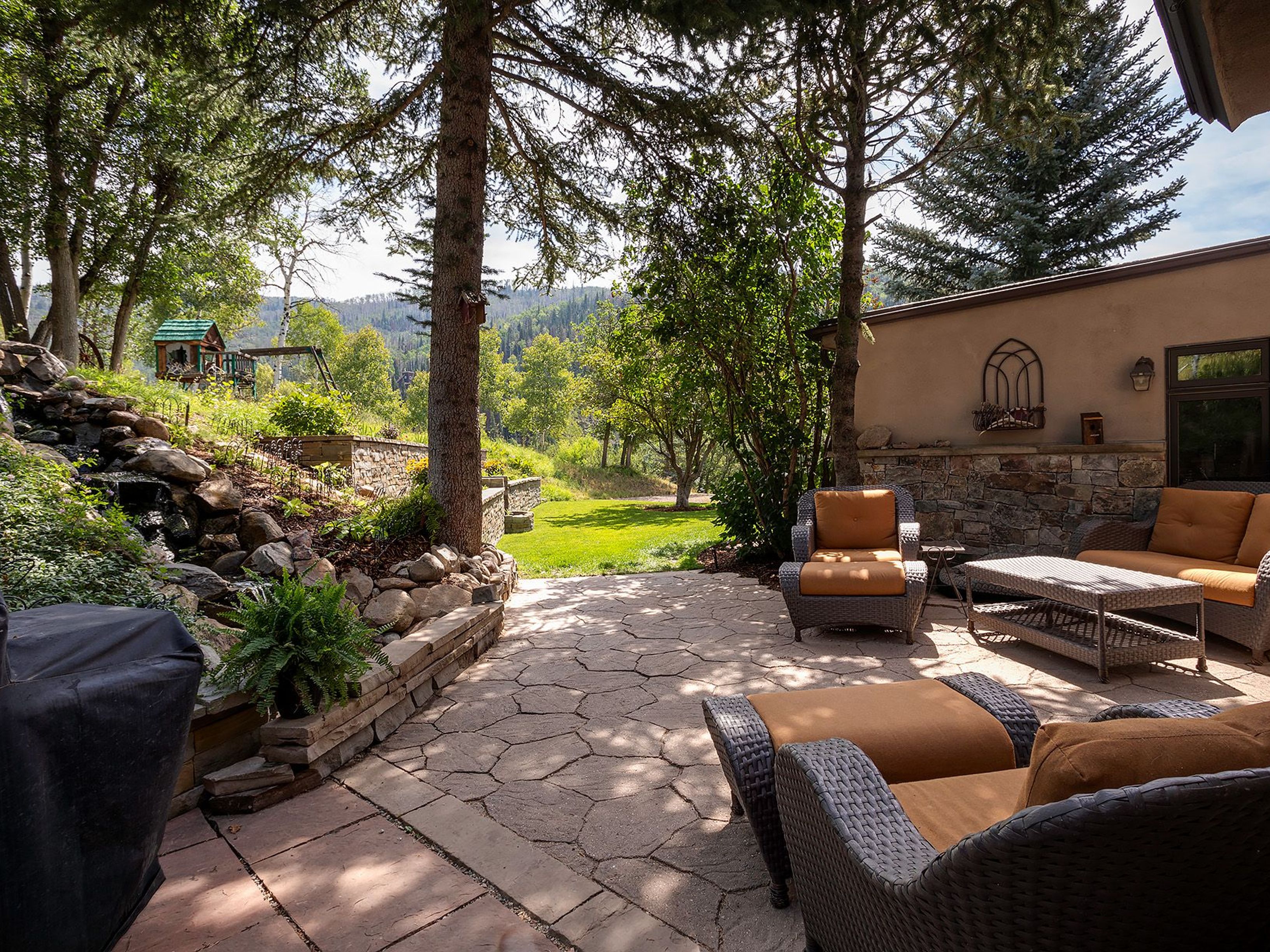 This image showcases a beautifully designed outdoor patio area, perfect for relaxation and entertaining. The patio features stone paving, comfortable wicker furniture with plush cushions, and a view of a lush green lawn and surrounding trees. A stone retaining wall with a small waterfall adds a touch of natural elegance to the space, creating a serene and inviting atmosphere.