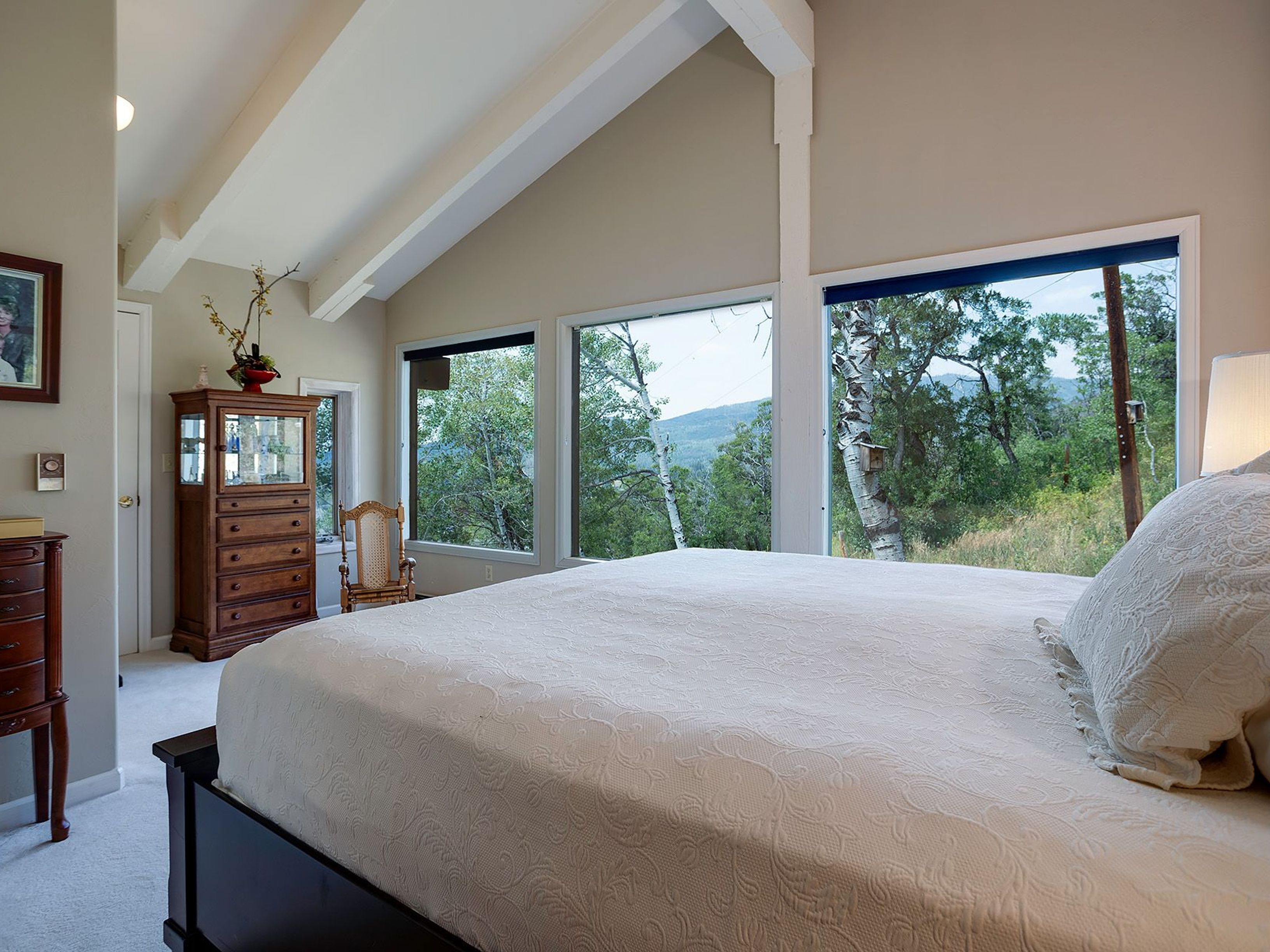 This primary bedroom features a large bed with a white patterned comforter, positioned to take advantage of the expansive windows offering views of the surrounding trees and mountains. The room has a vaulted ceiling with exposed beams, adding architectural interest, and is furnished with antique wooden dressers and a chair, creating a cozy and inviting atmosphere.