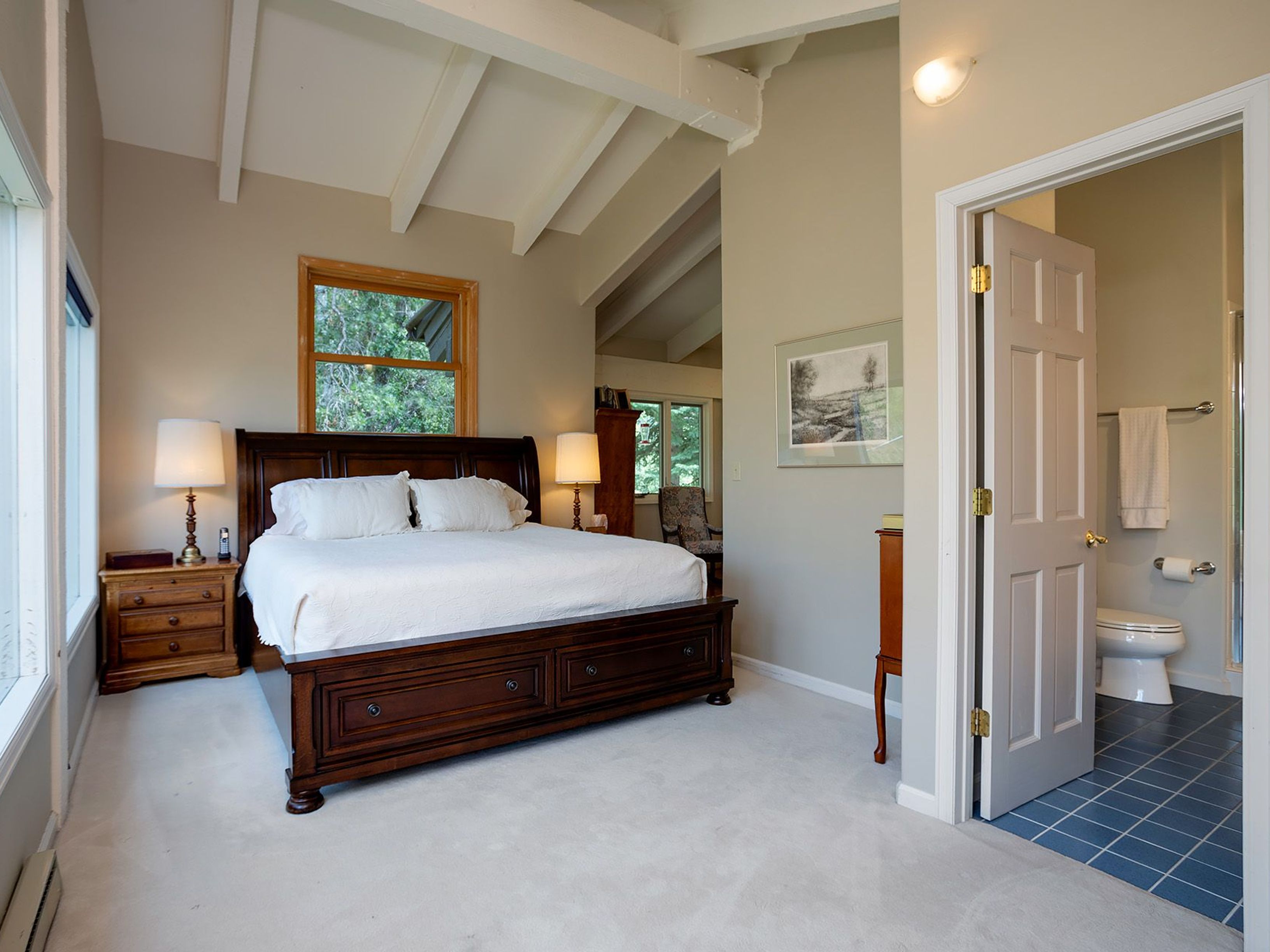 This is a primary bedroom featuring a large, dark wood bed with white linens, flanked by matching nightstands and lamps. The room has a vaulted ceiling with white beams and a window providing natural light. An open doorway leads to an ensuite bathroom with blue tile flooring, adding a touch of luxury and convenience.