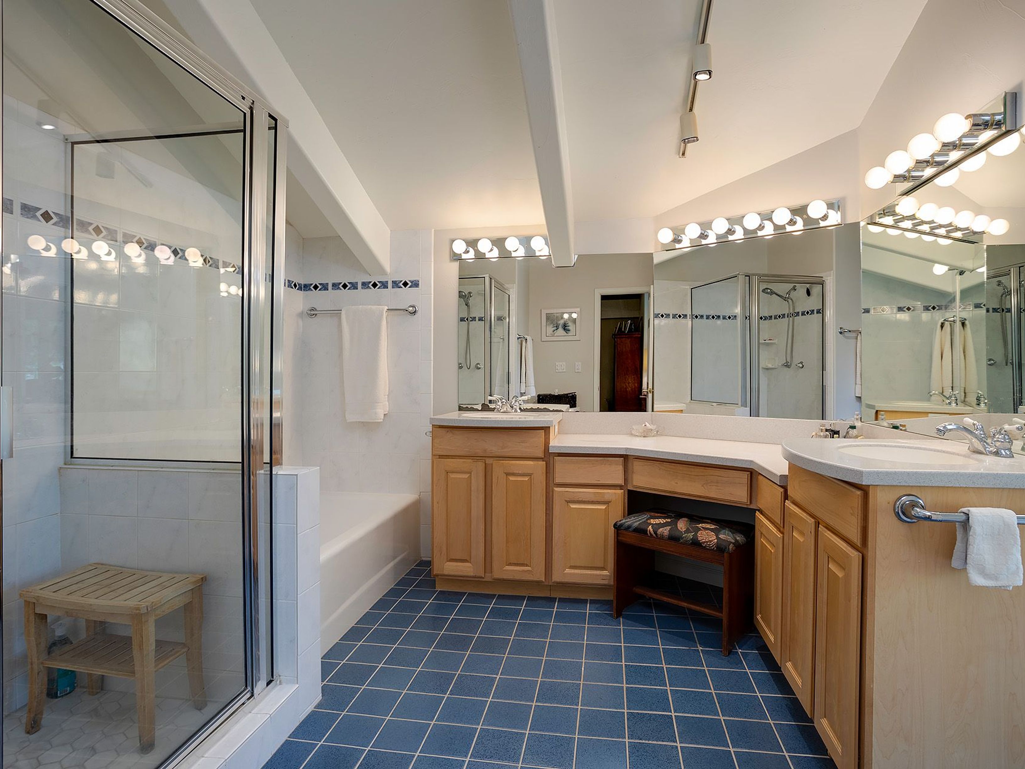 This is a well-lit primary bathroom featuring a double vanity with light wood cabinetry and a white countertop. A large mirror spans the length of the vanity, illuminated by multiple globe lights. The floor is tiled in a blue square pattern, and a glass-enclosed shower and tub are visible to the left, creating a clean and functional space.