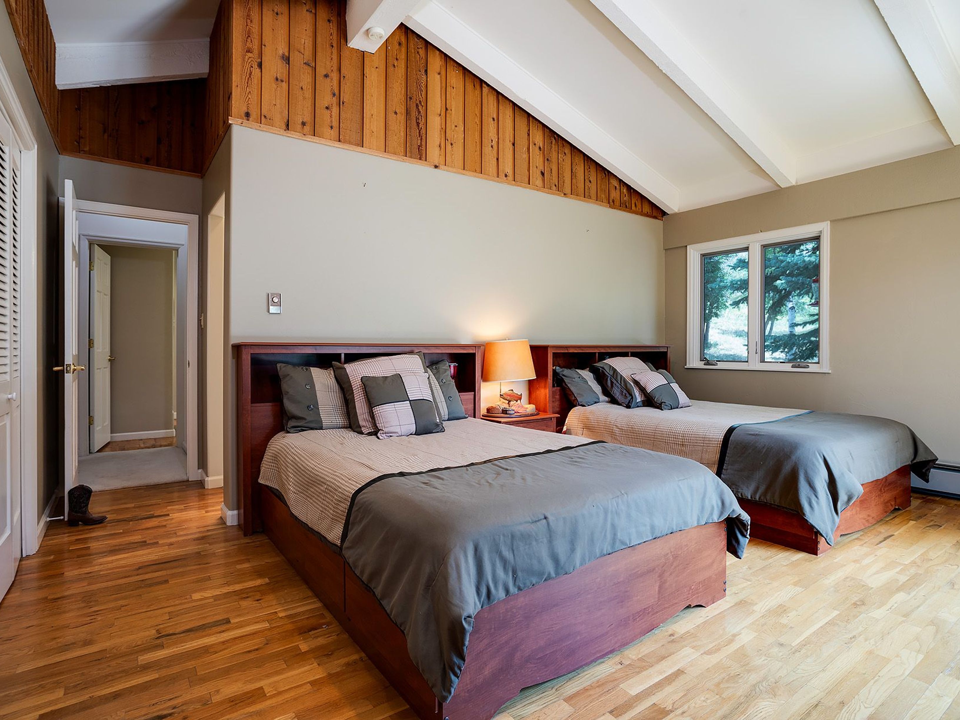 This is a bedroom featuring two twin beds with wooden frames and matching headboards. The room has hardwood floors, a window providing natural light, and a unique ceiling with wooden accents. The overall impression is cozy and inviting, suitable for guests or family members.