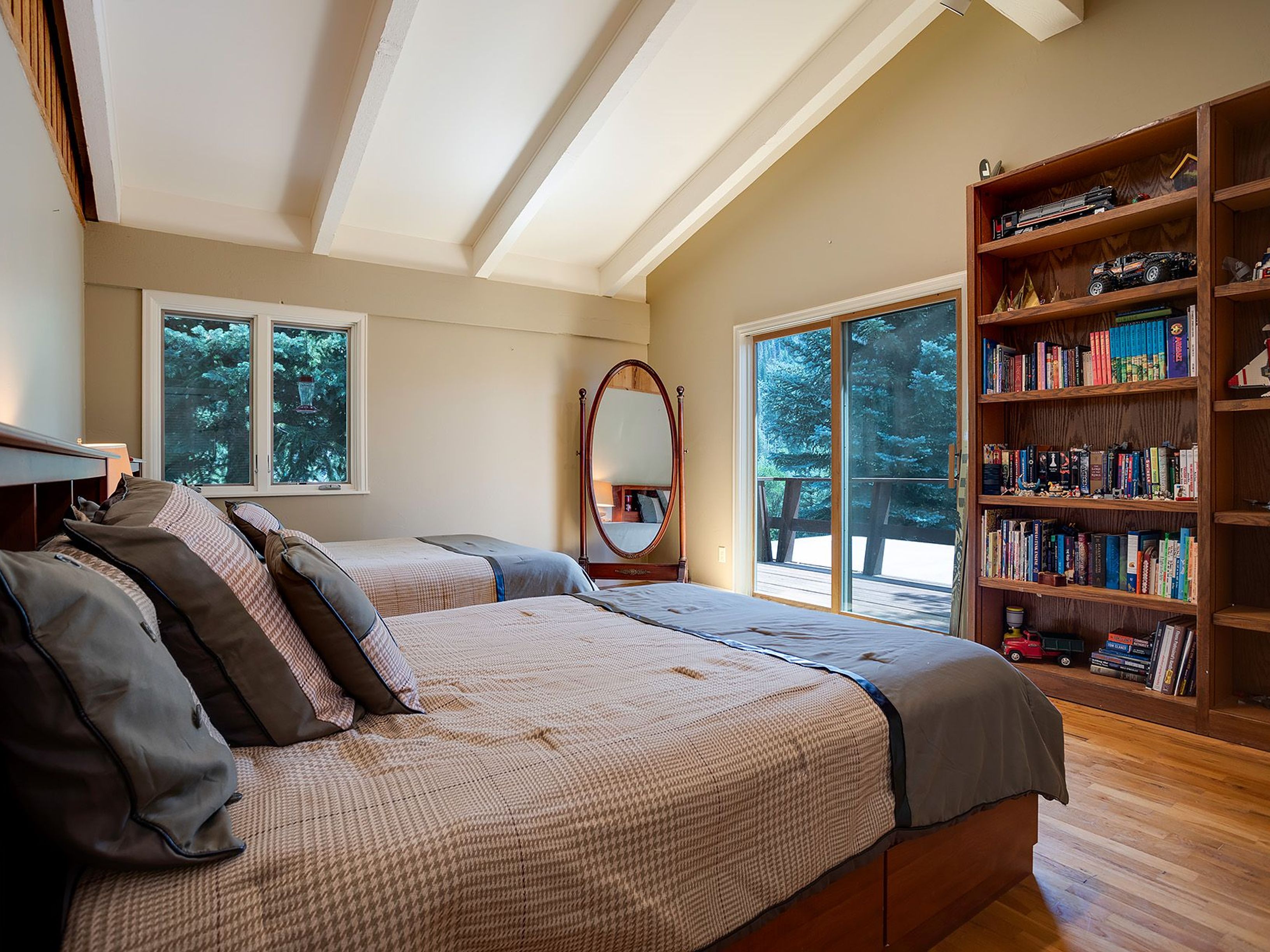 This is a cozy guest bedroom featuring two twin beds with patterned bedding and decorative pillows. A large oval mirror stands between the beds and a sliding glass door leads to an outdoor deck. A tall wooden bookshelf filled with books and decorative items adds character to the room, which is illuminated by natural light streaming through the windows and door.
