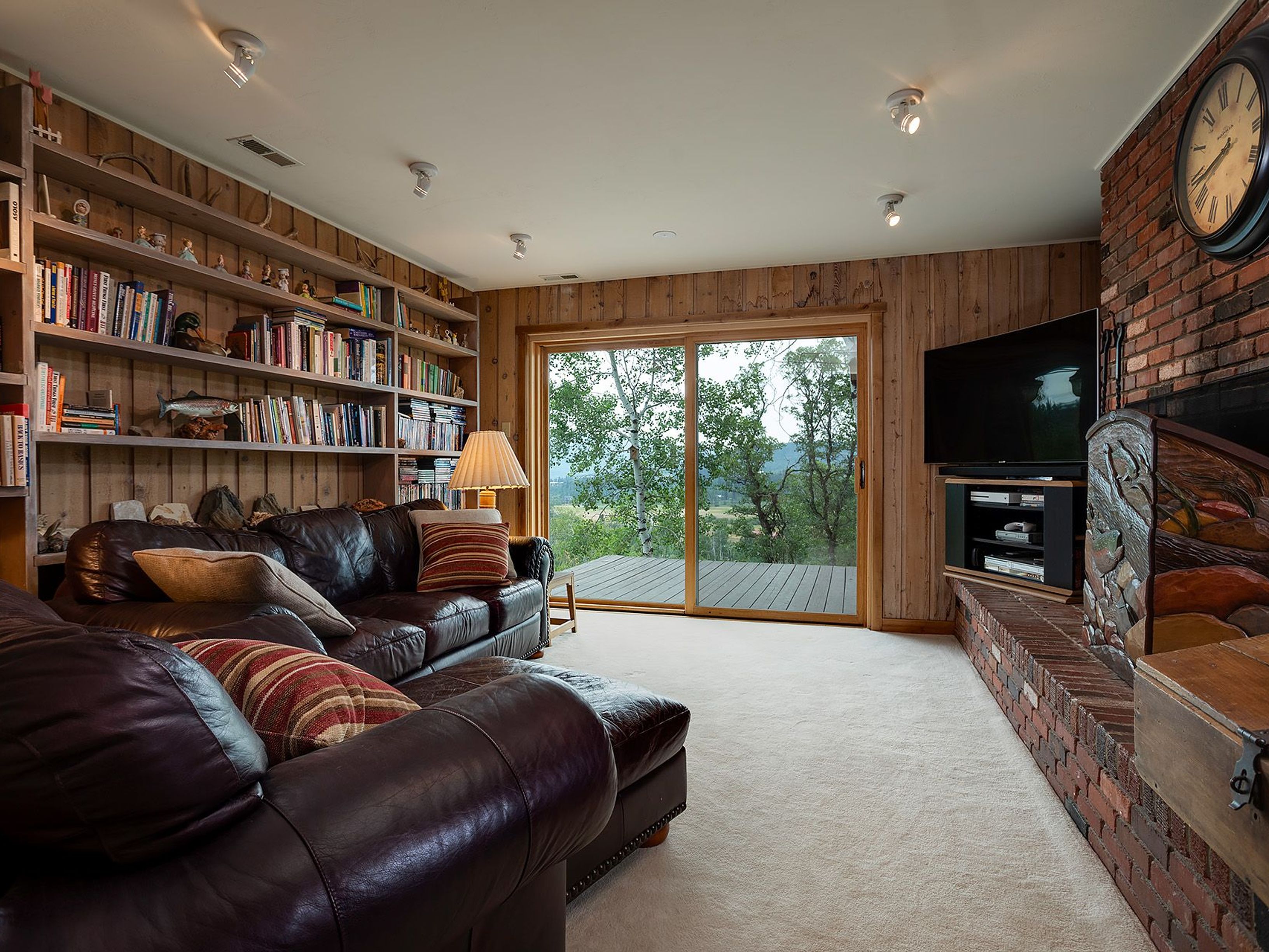 This inviting living room features a comfortable, dark leather sectional sofa with decorative pillows, a built-in bookshelf filled with books and decorative items, and a brick fireplace with a mounted television. A sliding glass door provides access to an outdoor deck and offers a view of the surrounding landscape. The room is carpeted and has wood-paneled walls, creating a warm and cozy atmosphere.