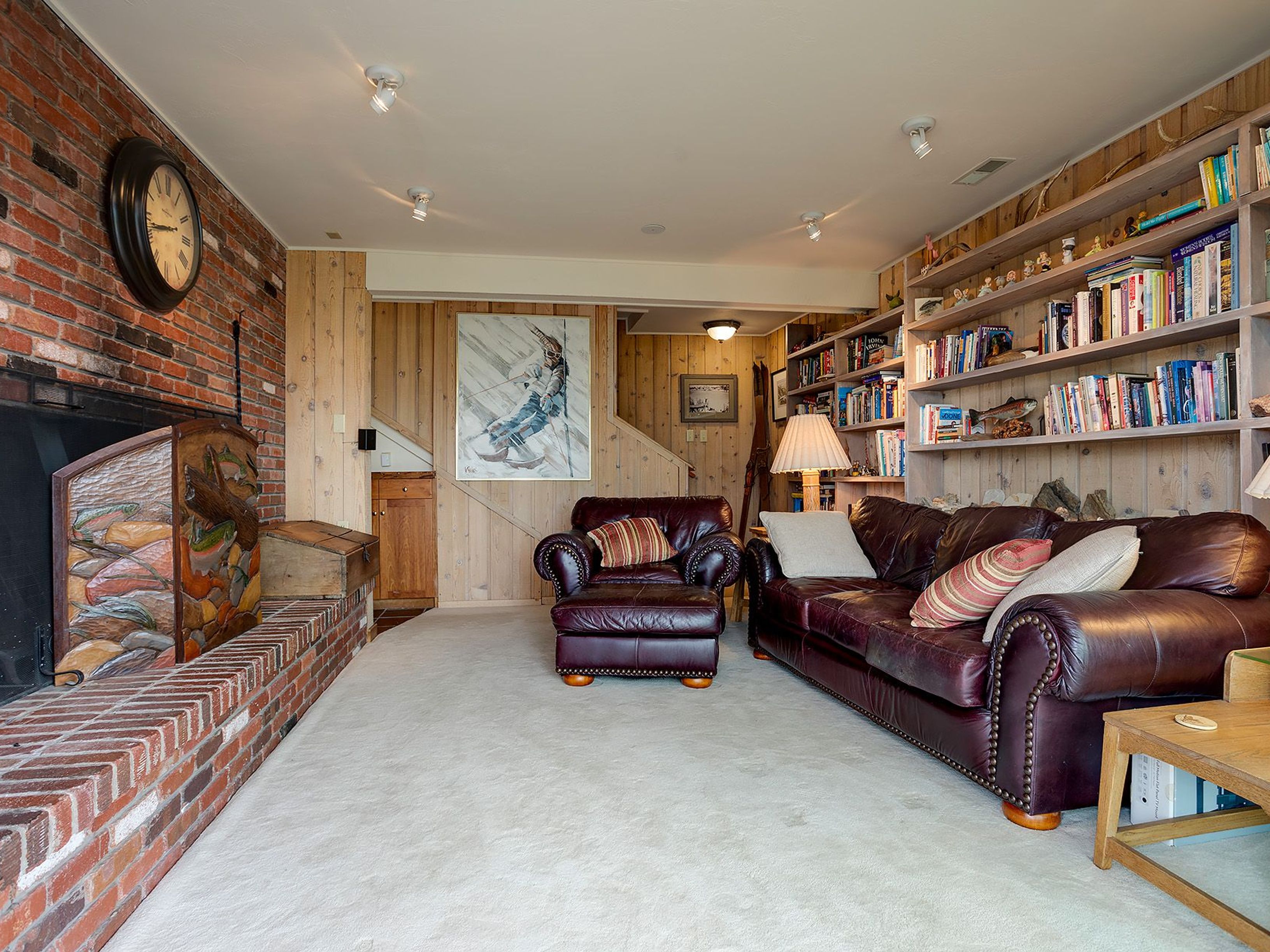 This inviting living room features a brick fireplace with a unique carved wood accent, complemented by wood-paneled walls and built-in bookshelves filled with books and decorative items. A comfortable leather sofa and armchair create a cozy seating area, illuminated by recessed lighting and a table lamp. The room exudes a warm, rustic charm perfect for relaxation and entertainment.
