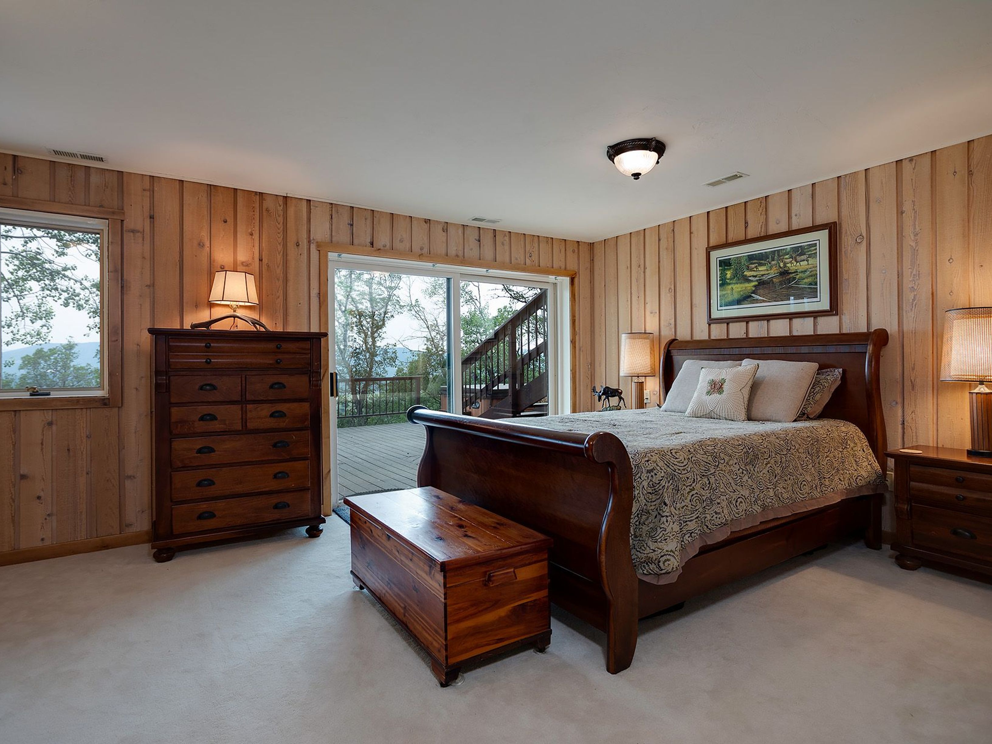 This is a cozy primary bedroom featuring wood-paneled walls and a traditional sleigh bed with neutral bedding. A wooden chest sits at the foot of the bed, and matching nightstands and dressers flank the sides. A sliding glass door leads to an outdoor deck, offering a view of the surrounding landscape.
