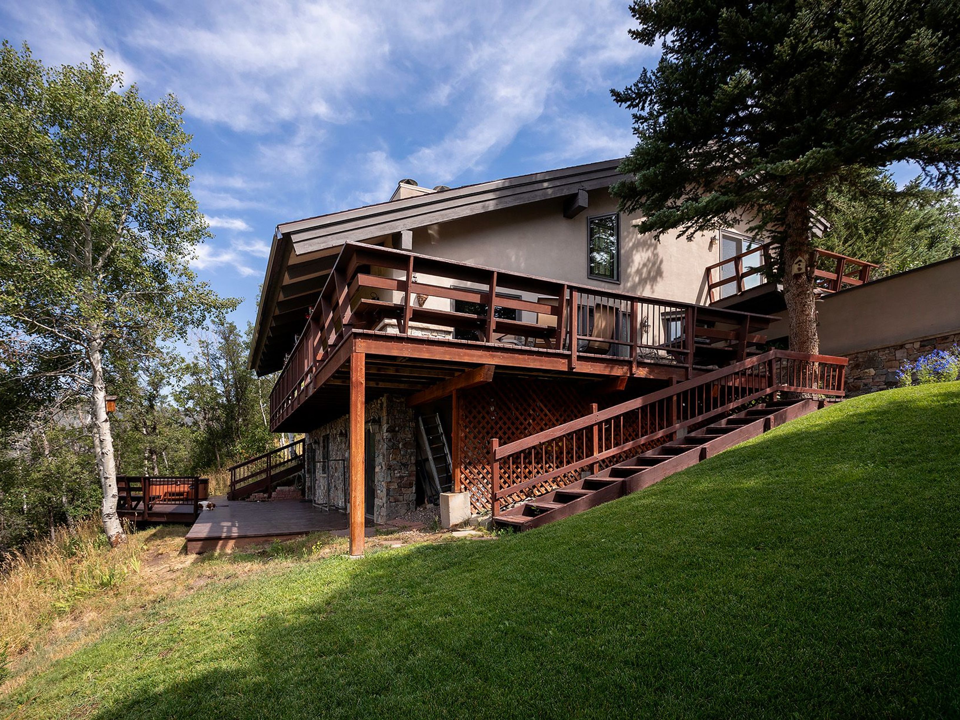 This image showcases the rear exterior of a multi-level home with a prominent wooden deck and staircase leading down to the yard. The house features a combination of stone and stucco siding, blending with the natural landscape. Lush green grass covers the yard, creating a serene and inviting outdoor space.