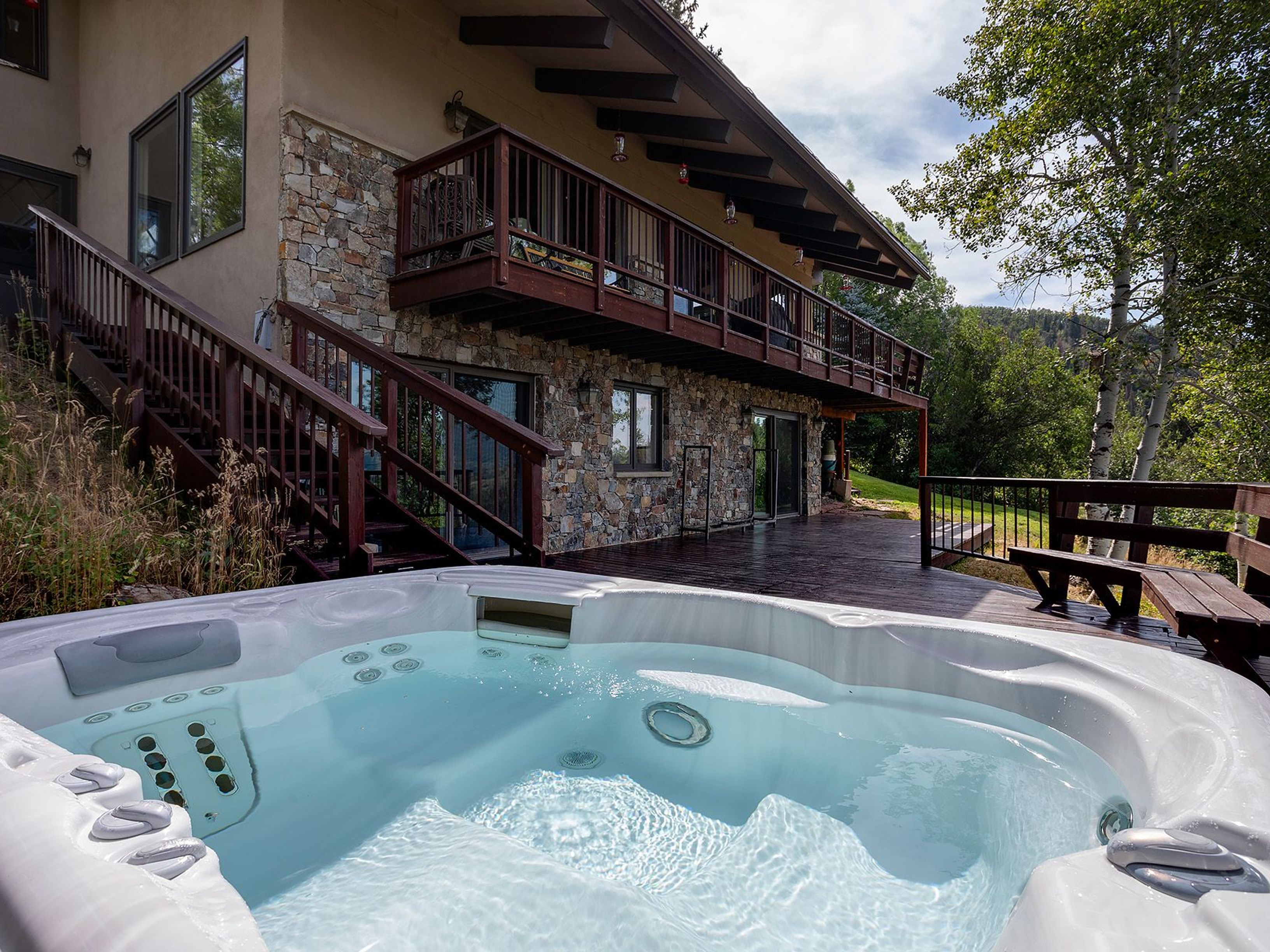 This image showcases a luxurious outdoor spa area, featuring a large hot tub with crystal-clear water. The spa is situated on a spacious wooden deck, adjacent to a multi-level house with stone accents and a wooden balcony. The setting is surrounded by lush greenery, creating a private and serene atmosphere.
