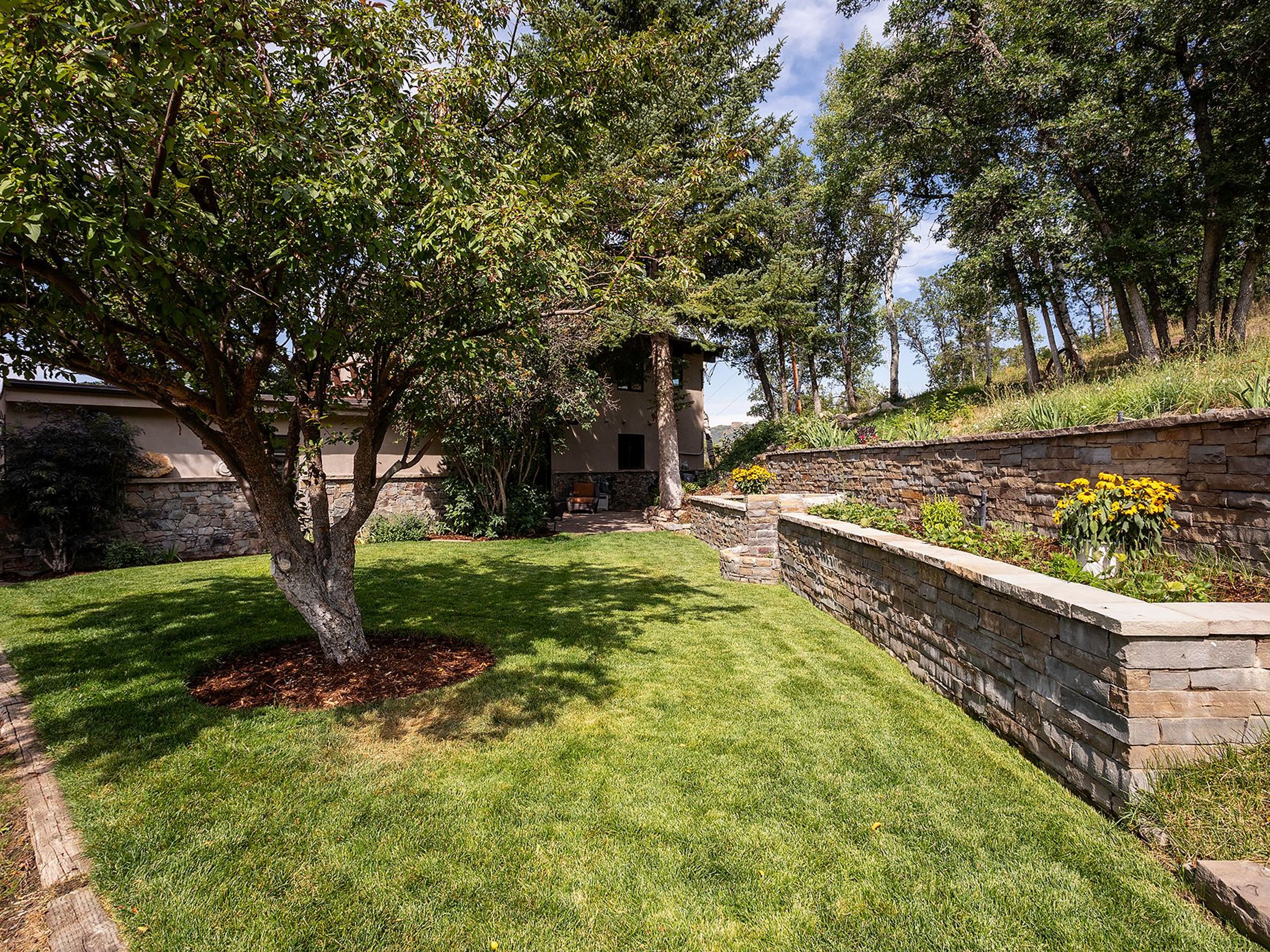 This image showcases a beautifully landscaped backyard featuring a lush green lawn, mature trees providing shade, and a stone retaining wall with planted flowers. The yard is well-maintained and offers a serene outdoor space, enhancing the property's appeal. The perspective is from ground level, capturing the depth and tranquility of the garden.