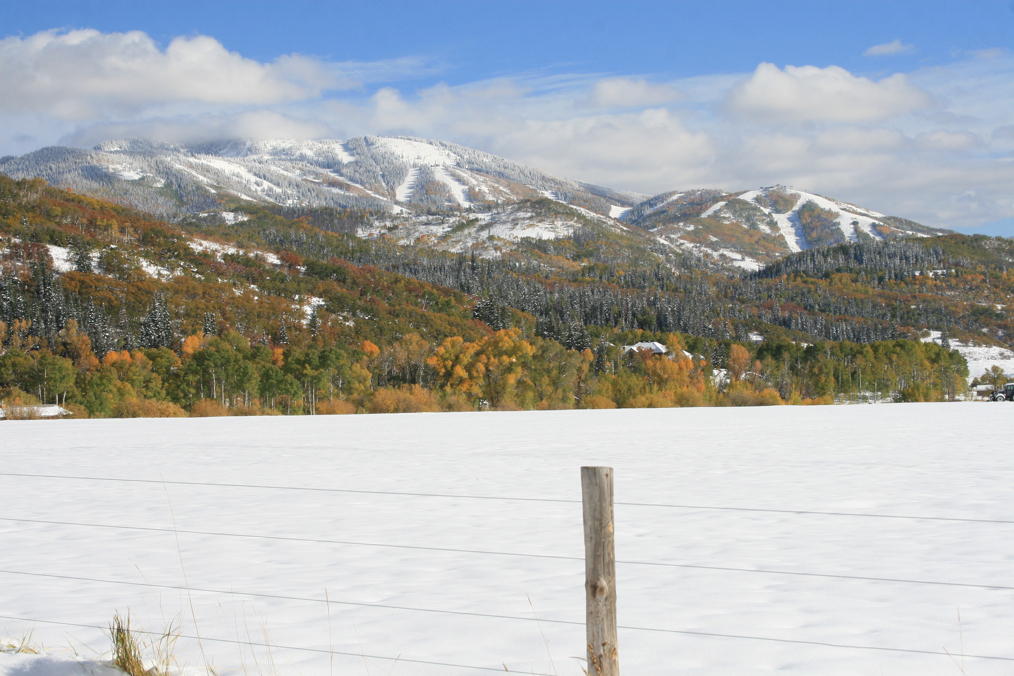 The image showcases a vast, snow-covered yard or field, framed by a wooden fence post in the foreground. In the background, a picturesque mountain range is visible, partially covered in snow and interspersed with trees displaying autumn foliage. The scene evokes a sense of serene, natural beauty, highlighting the property's expansive outdoor space and scenic views.