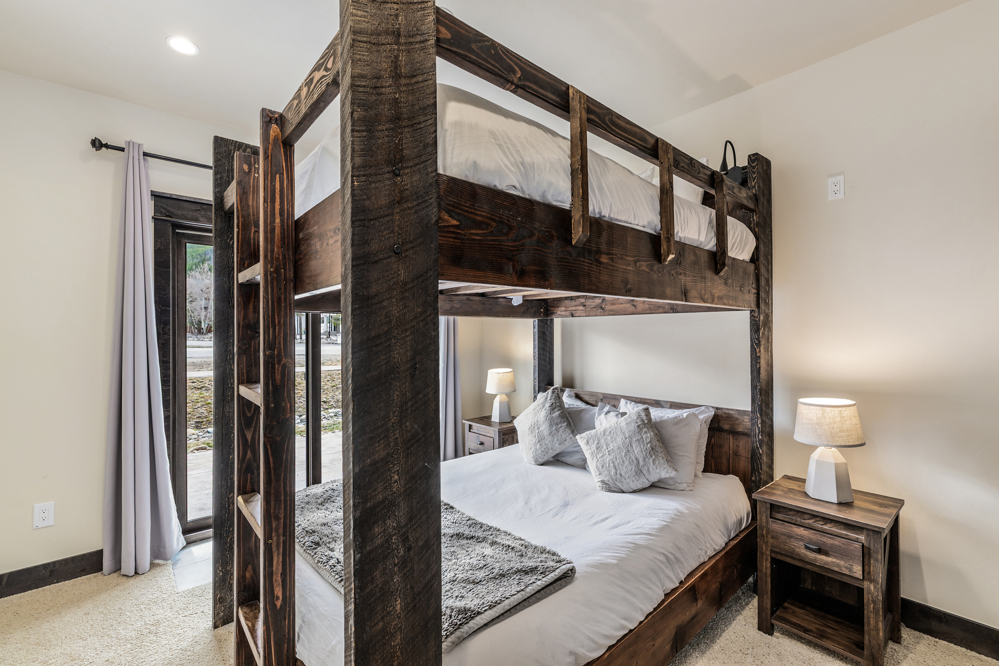 This cozy bedroom features a rustic, dark-stained wooden bunk bed structure that frames a lower queen-sized bed with plush white bedding and decorative pillows. The room is accented by matching wooden nightstands with modern lamps, creating a warm, cabin-inspired aesthetic. A sliding glass door to the left provides natural light and a view of the outdoors, while the neutral walls and carpet maintain a clean, inviting atmosphere.