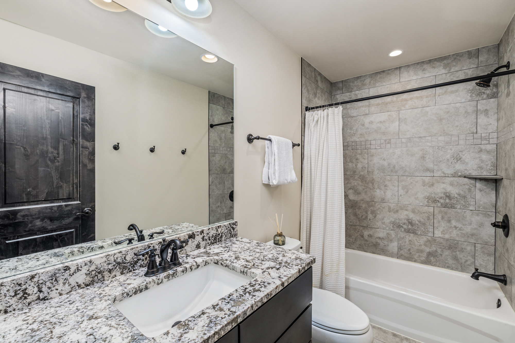 This modern bathroom features a granite vanity countertop with a dark wood cabinet and a sleek, dark-finished faucet. The space includes a shower-tub combination with grey stone-look tile surround and a matching dark shower head and curtain rod. The neutral color palette and clean lines create a contemporary and functional aesthetic.