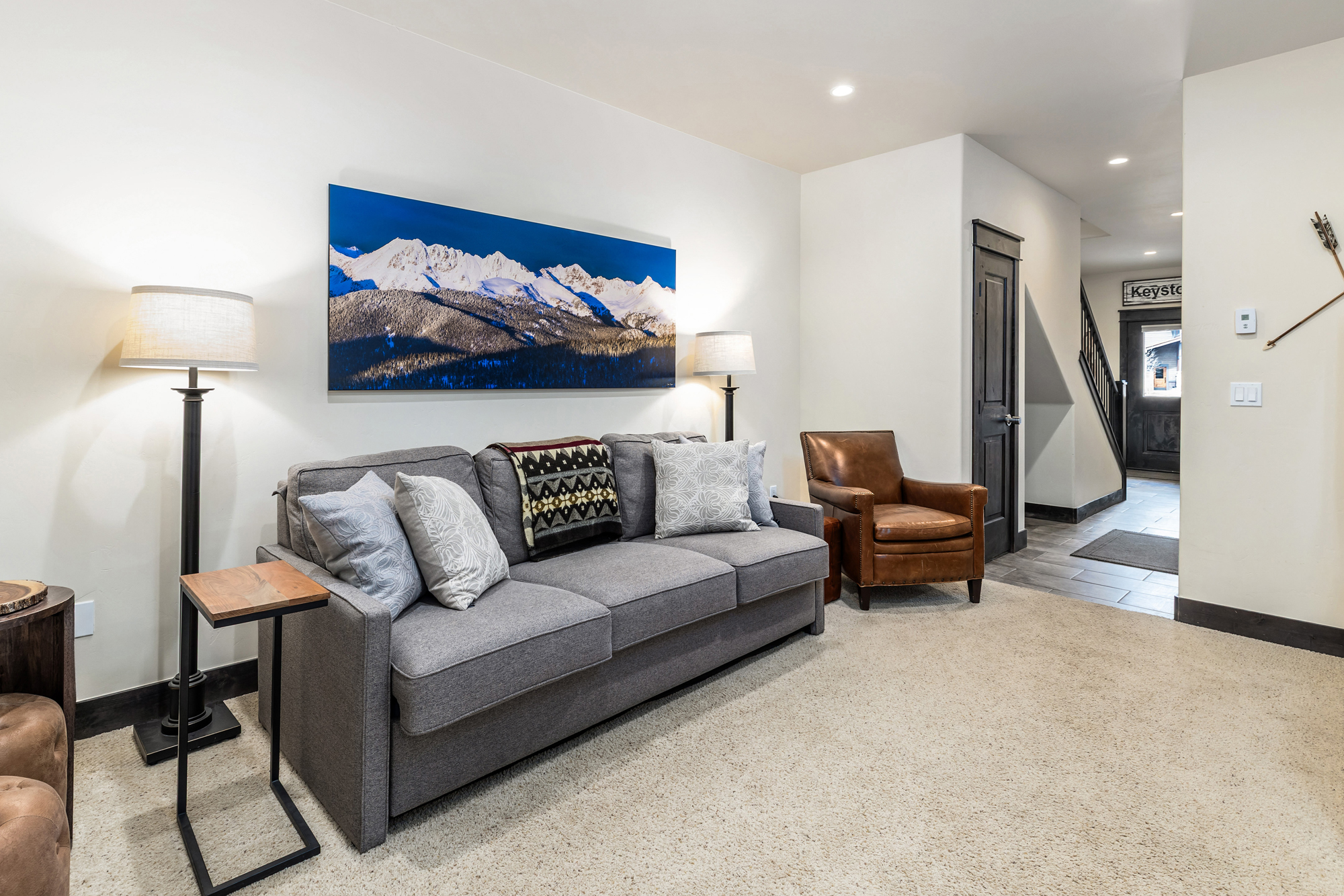 This cozy living room features a comfortable grey sofa adorned with patterned throw pillows and a rustic mountain-themed blanket, centered beneath a large, vibrant landscape print of snow-capped mountains. To the right, a classic brown leather armchair adds a touch of warmth, while a sleek side table and floor lamps provide functional lighting. The space is finished with neutral carpeting and leads into a hallway, creating an inviting and well-appointed atmosphere.