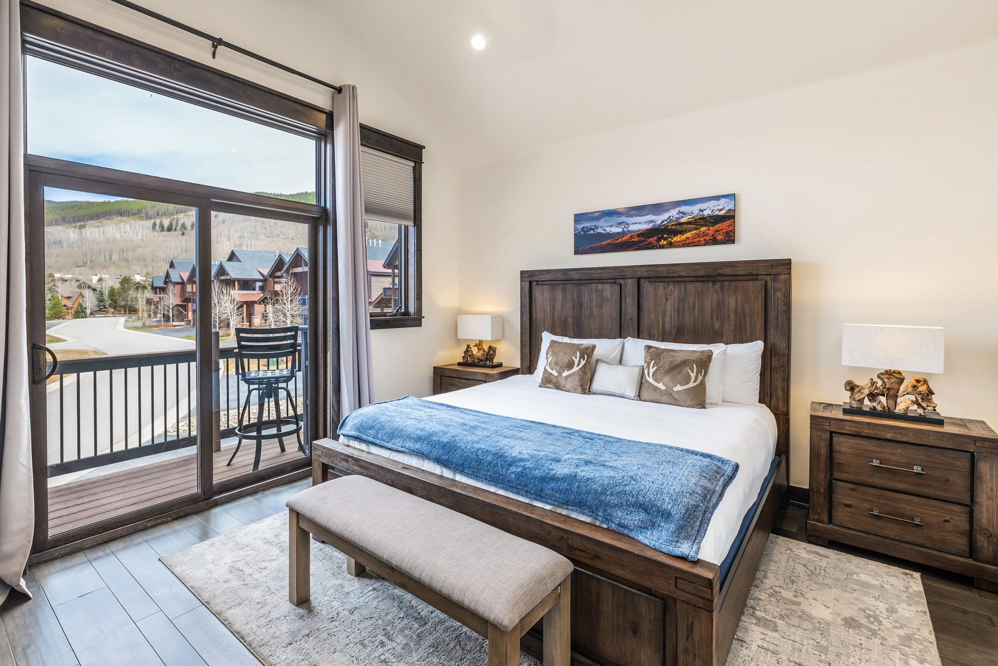 This primary bedroom features a rustic, mountain-inspired aesthetic with a large wooden bed frame, a cozy blue throw blanket, and decorative antler-themed pillows. A sliding glass door provides abundant natural light and direct access to a private balcony overlooking a scenic neighborhood. The room is accented by warm bedside lamps and a panoramic landscape painting above the headboard, creating a serene and inviting atmosphere.
