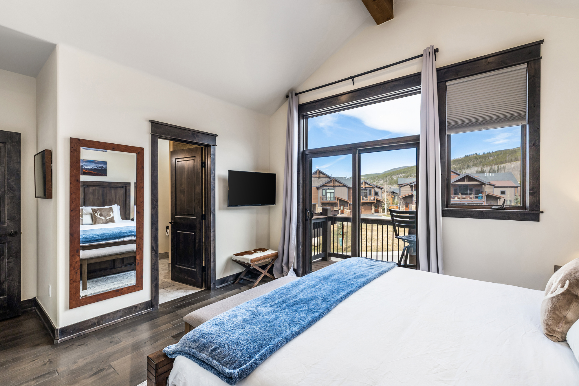 This primary bedroom features a rustic-modern aesthetic with dark wood flooring, trim, and a matching framed mirror reflecting the bed. A large glass sliding door provides abundant natural light and a scenic view of the surrounding mountain community, while a blue throw blanket adds a pop of color to the neutral bedding. The room is well-appointed with a wall-mounted television and a small stool, creating a cozy and inviting atmosphere.