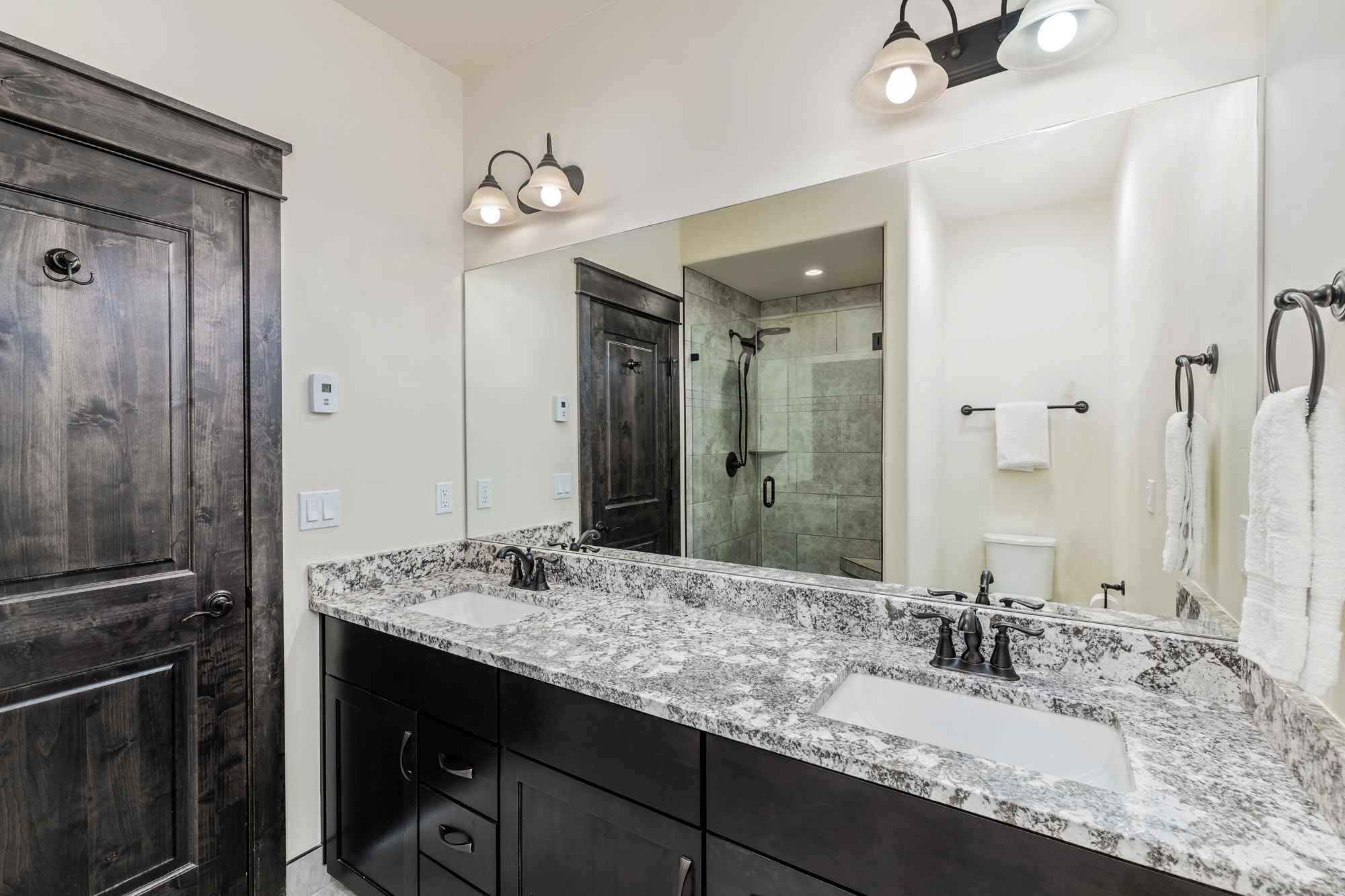 This primary bathroom features a double vanity with dark wood cabinetry and a speckled granite countertop, complemented by dual under-mount sinks and oil-rubbed bronze fixtures. A large, frameless mirror spans the wall above the vanity, reflecting the adjacent walk-in shower and toilet area. The space is illuminated by two stylish wall-mounted light fixtures, creating a clean, functional, and modern aesthetic.