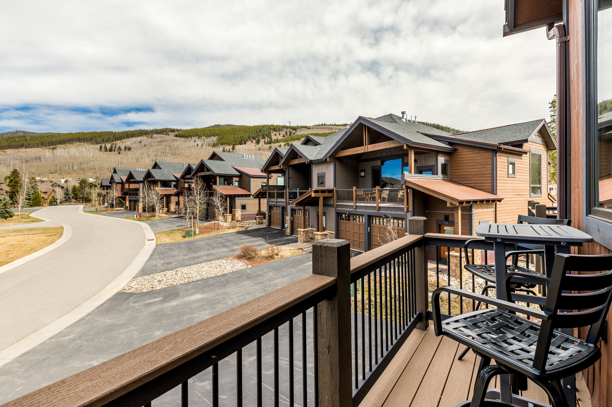 This image captures a scenic view from a wooden balcony, featuring a bistro set overlooking a row of modern mountain-style townhomes. The architecture showcases a blend of wood siding, stone accents, and dark trim, set against a backdrop of rolling hills and a cloudy sky. The perspective provides a clear, elevated vantage point of the neighborhood's layout and the surrounding natural landscape.