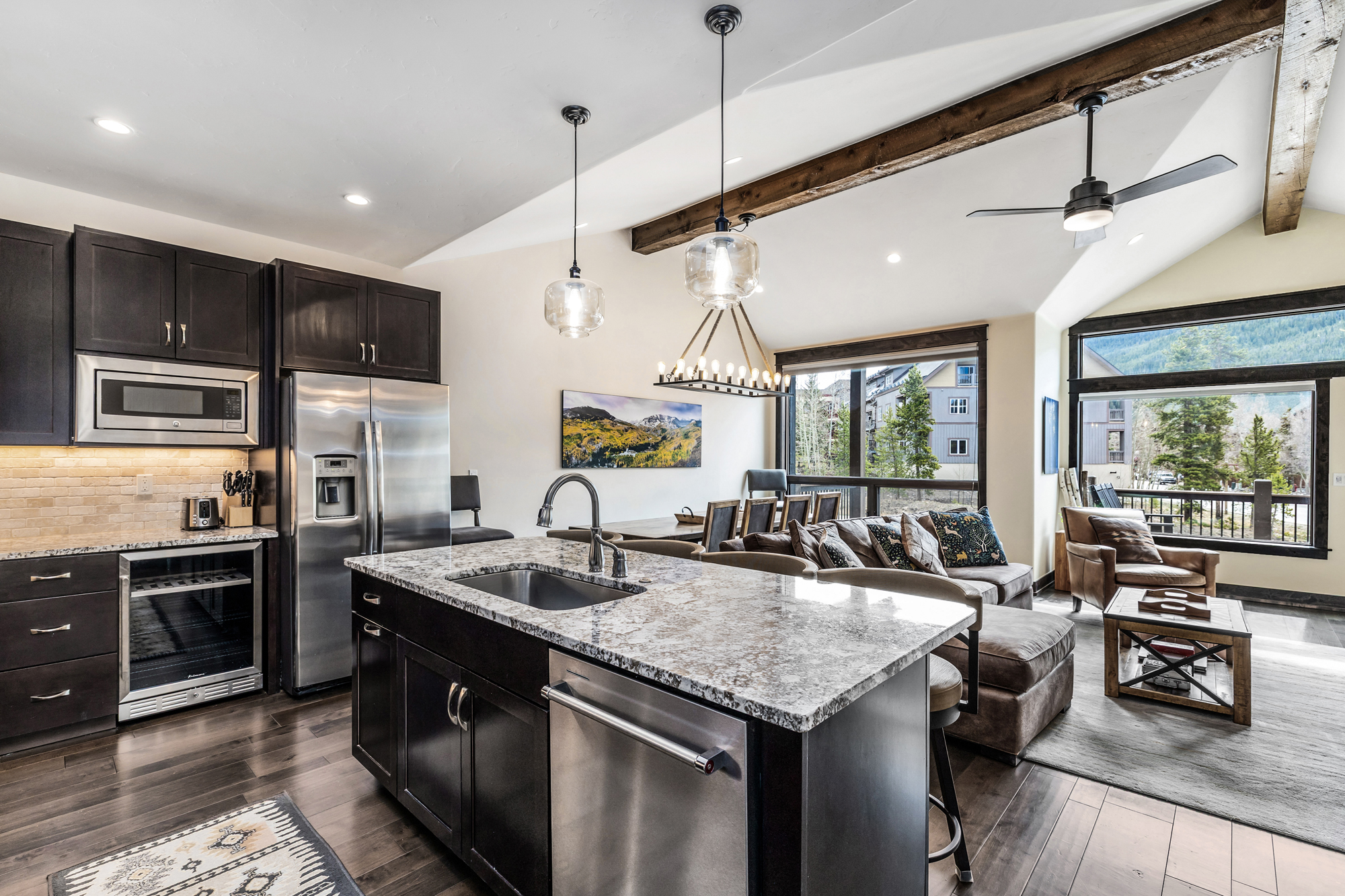 This open-concept kitchen features dark wood cabinetry, granite countertops, and high-end stainless steel appliances, including a built-in wine cooler. The space flows seamlessly into a dining and living area, highlighted by vaulted ceilings with exposed wooden beams and large windows that offer scenic mountain views. The warm, rustic-modern aesthetic is enhanced by stylish pendant lighting and a spacious kitchen island, creating an inviting atmosphere for entertaining.