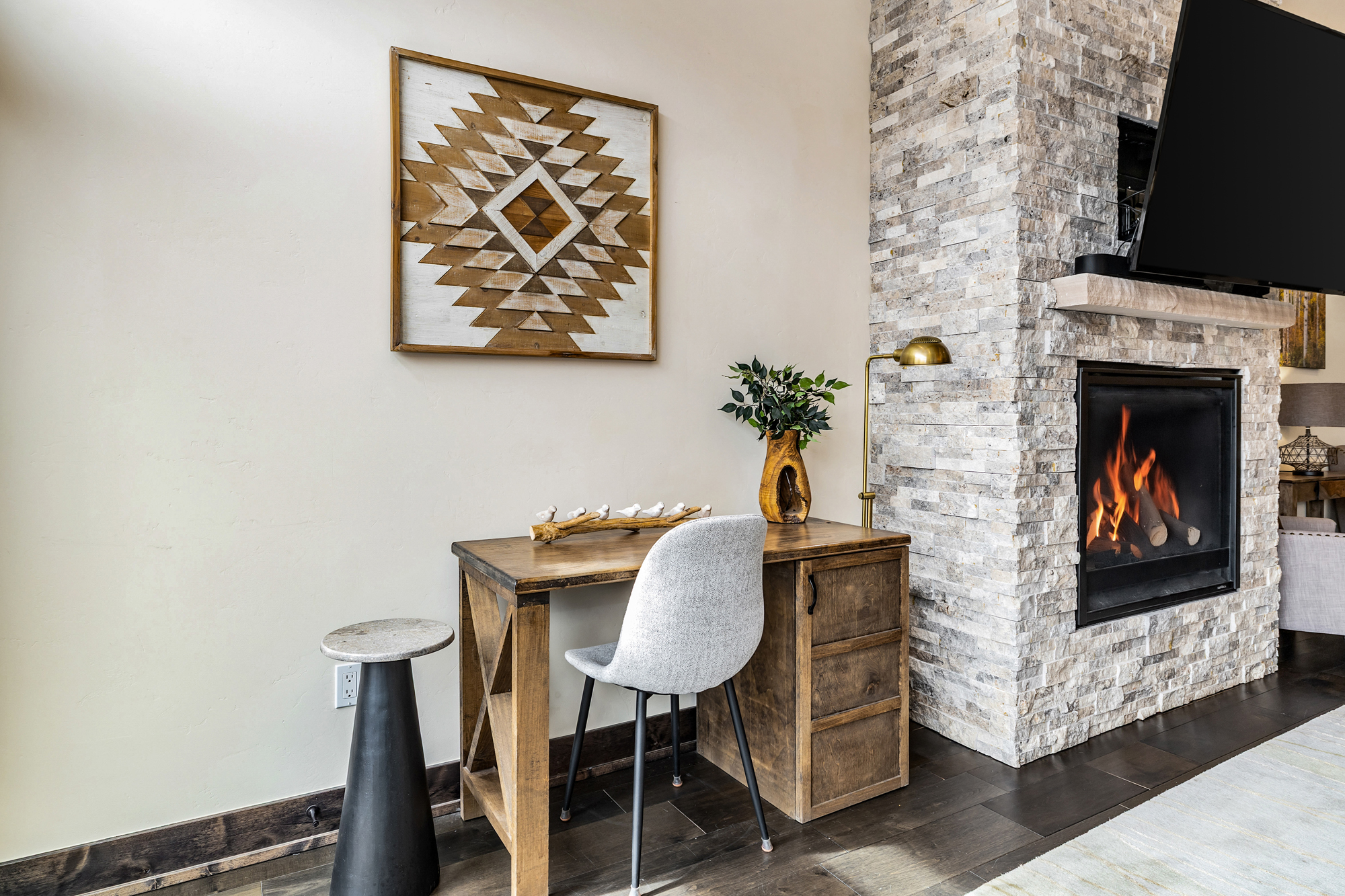 This cozy home office nook features a rustic wooden desk paired with a modern grey upholstered chair, set against a neutral wall adorned with a geometric wood art piece. To the right, a striking floor-to-ceiling stone fireplace adds warmth and texture, while a sleek black pedestal table stands to the left. The space blends rustic charm with contemporary design, creating an inviting and functional workspace.