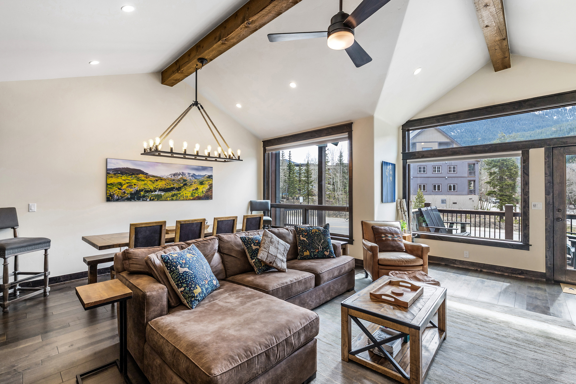 This spacious, open-concept living area features a vaulted ceiling with exposed wooden beams and a large, comfortable brown sectional sofa. A rustic wooden coffee table sits on a neutral area rug, while a dining table with a unique rope-suspended chandelier is positioned behind the sofa. Large windows provide ample natural light and scenic views of the surrounding mountain landscape, creating a warm and inviting mountain-lodge atmosphere.