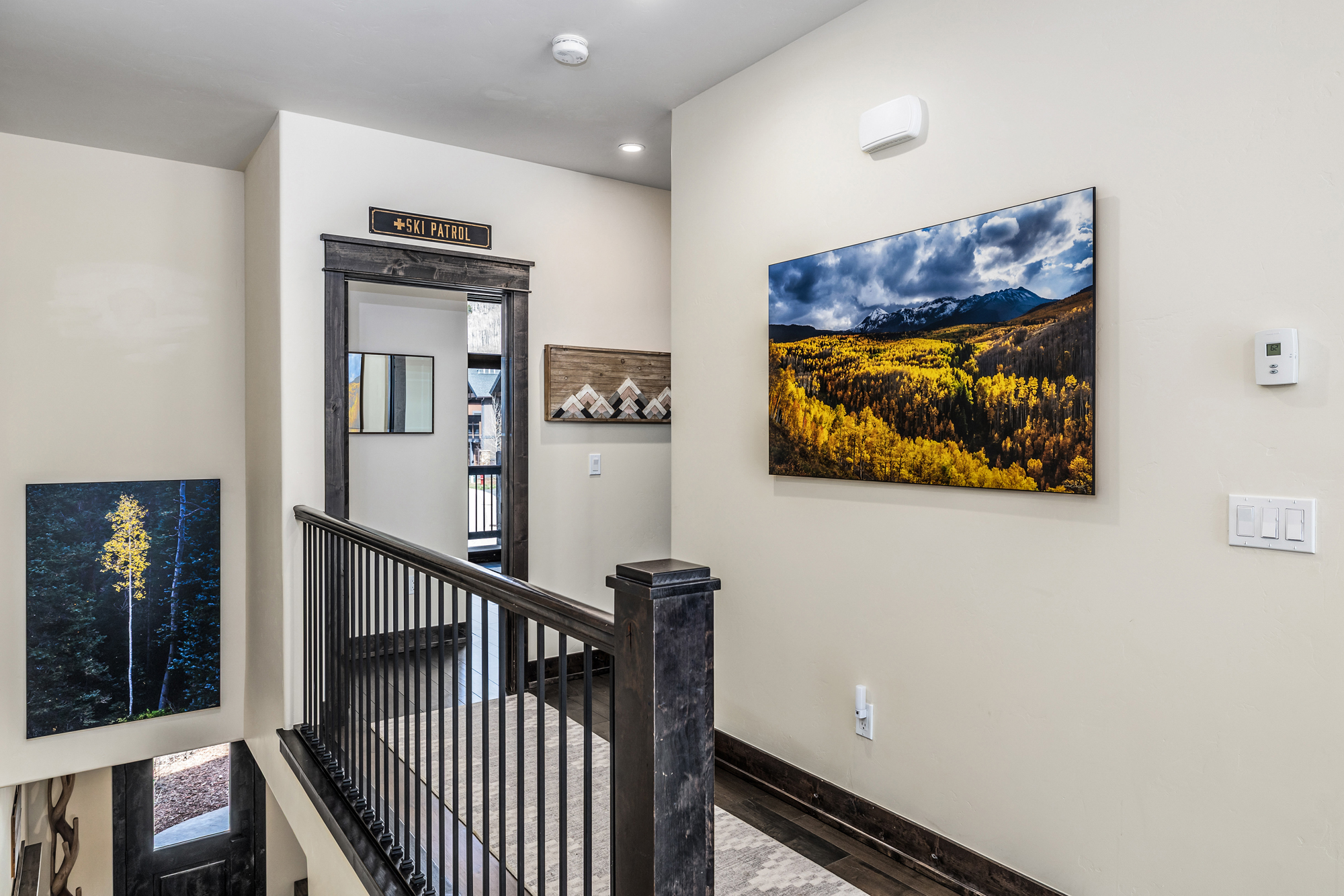 This interior hallway features a rustic, mountain-inspired aesthetic with dark wood trim, a metal railing, and nature-themed artwork. The space is bright and welcoming, highlighted by a 'Ski Patrol' sign above the doorway and a large landscape print of an autumn forest. The perspective looks down a short corridor toward a doorway, emphasizing the home's cozy, lodge-like character.