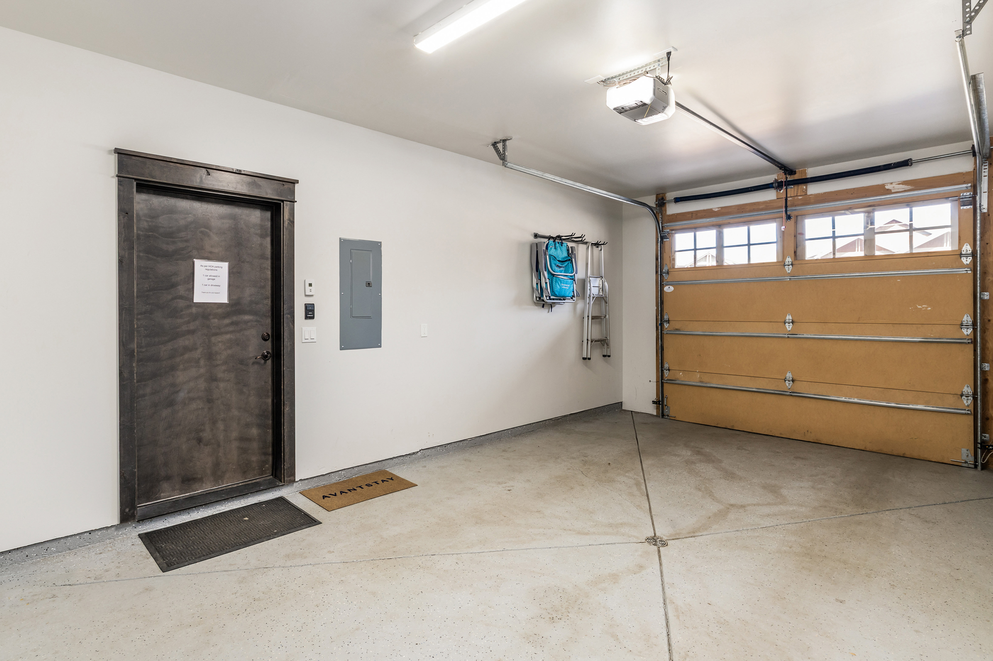 This image showcases a clean, well-maintained garage interior with a concrete floor and a prominent wooden garage door. The space features a side entry door with a dark frame, a wall-mounted electrical panel, and convenient storage hooks holding a folding chair and a small ladder. The perspective is wide-angle, capturing the functional layout and neutral aesthetic of the space.