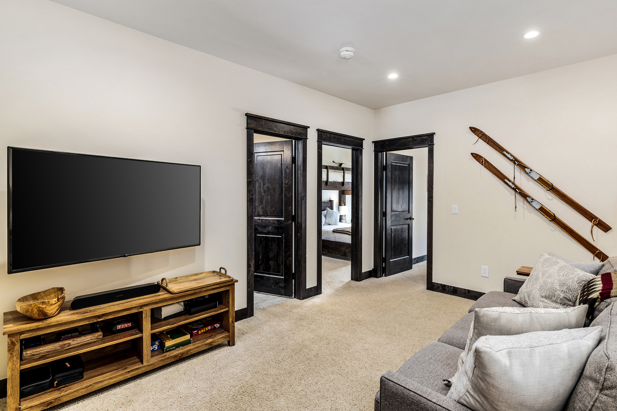 This cozy living room features a rustic aesthetic with dark wood trim, a wall-mounted television, and a wooden media console. A comfortable grey sofa sits to the right, accented by decorative pillows, while vintage wooden skis mounted on the wall add a charming mountain-lodge touch. The room provides a clear view into an adjacent bedroom, emphasizing a warm and inviting atmosphere.