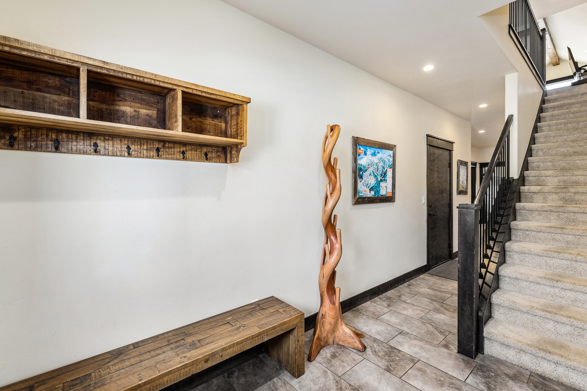 This inviting entryway features a rustic wooden bench and matching wall-mounted coat rack, complemented by a unique, sculptural wooden coat stand. The space leads to a carpeted staircase with a dark metal railing, set against a neutral color palette and tiled flooring. The overall aesthetic is warm and mountain-inspired, creating a welcoming transition area within the home.