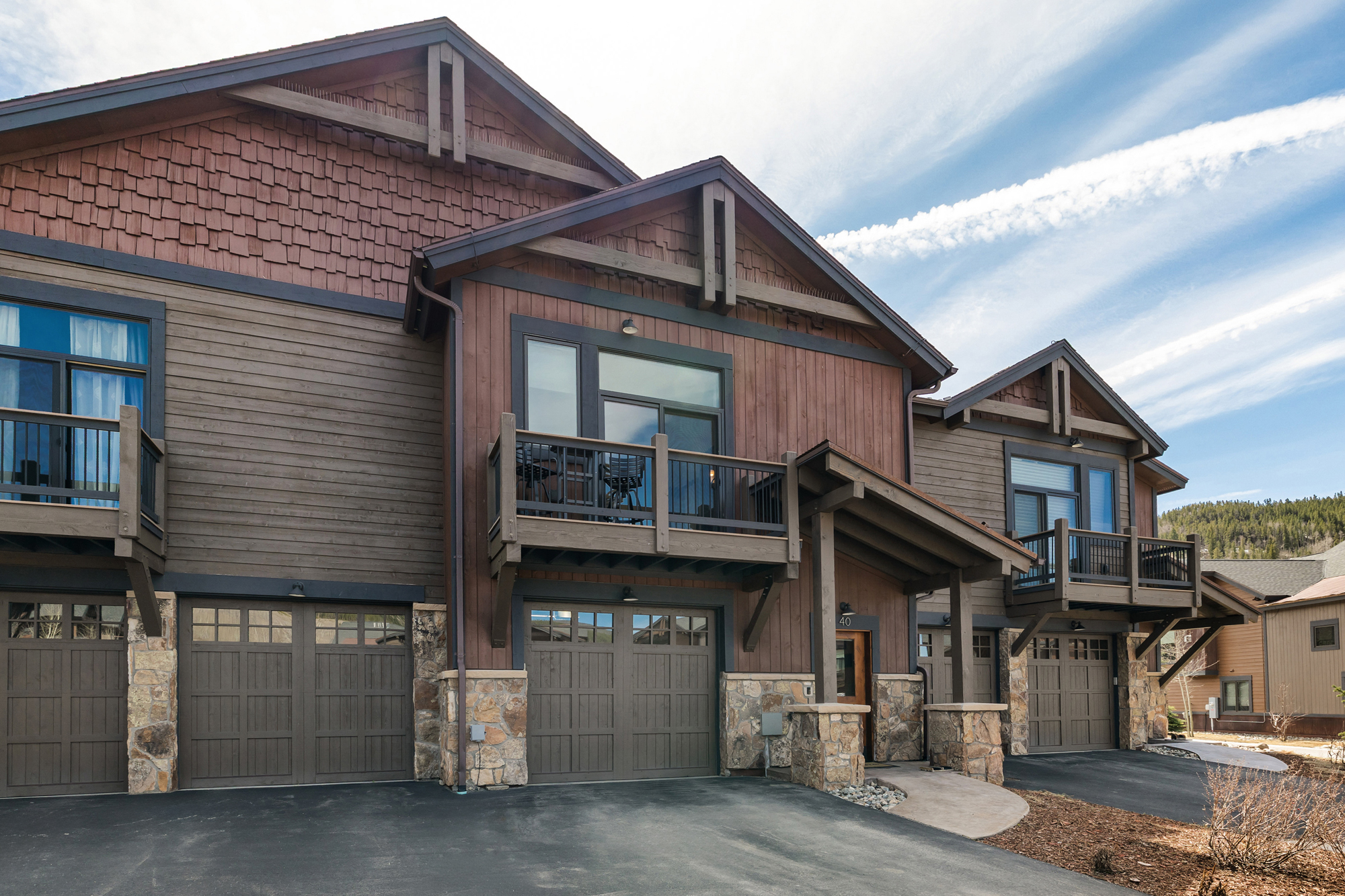 This image captures the front exterior of a multi-unit mountain-style townhouse complex, showcasing a blend of rustic stone, vertical wood siding, and shingle accents. The structure features multiple garage bays, elevated balconies with black railings, and gabled rooflines that evoke a cozy, upscale lodge aesthetic. The perspective is a wide-angle, eye-level shot that highlights the architectural depth and the inviting, well-maintained driveway leading to the entrances.