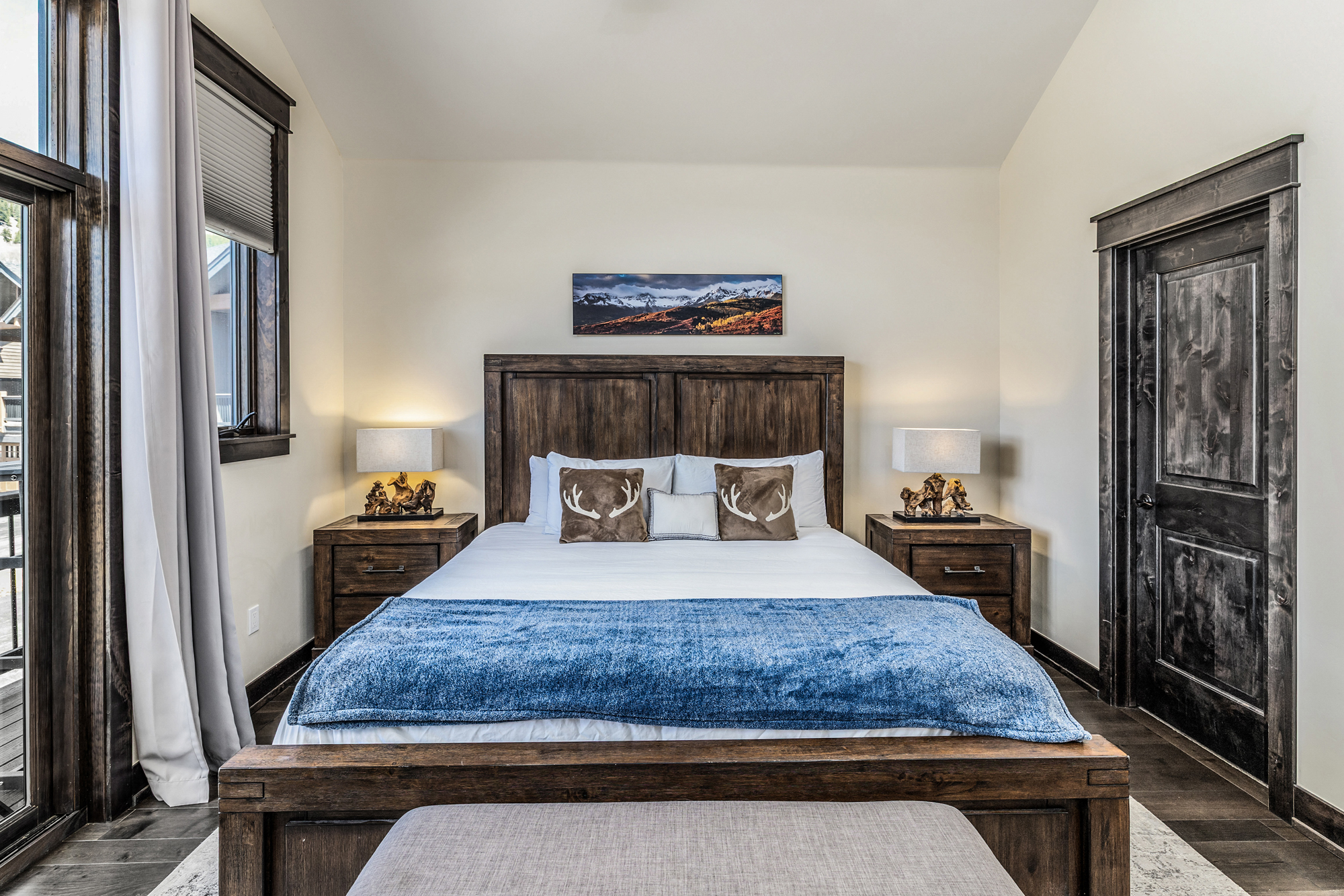 This primary bedroom features a rustic, mountain-inspired aesthetic with a substantial dark wood bed frame and matching nightstands. The room is anchored by a plush blue throw blanket, decorative antler-themed pillows, and a landscape painting above the headboard, all set against a neutral backdrop. The perspective is a centered, eye-level shot that highlights the cozy, high-end lodge atmosphere.