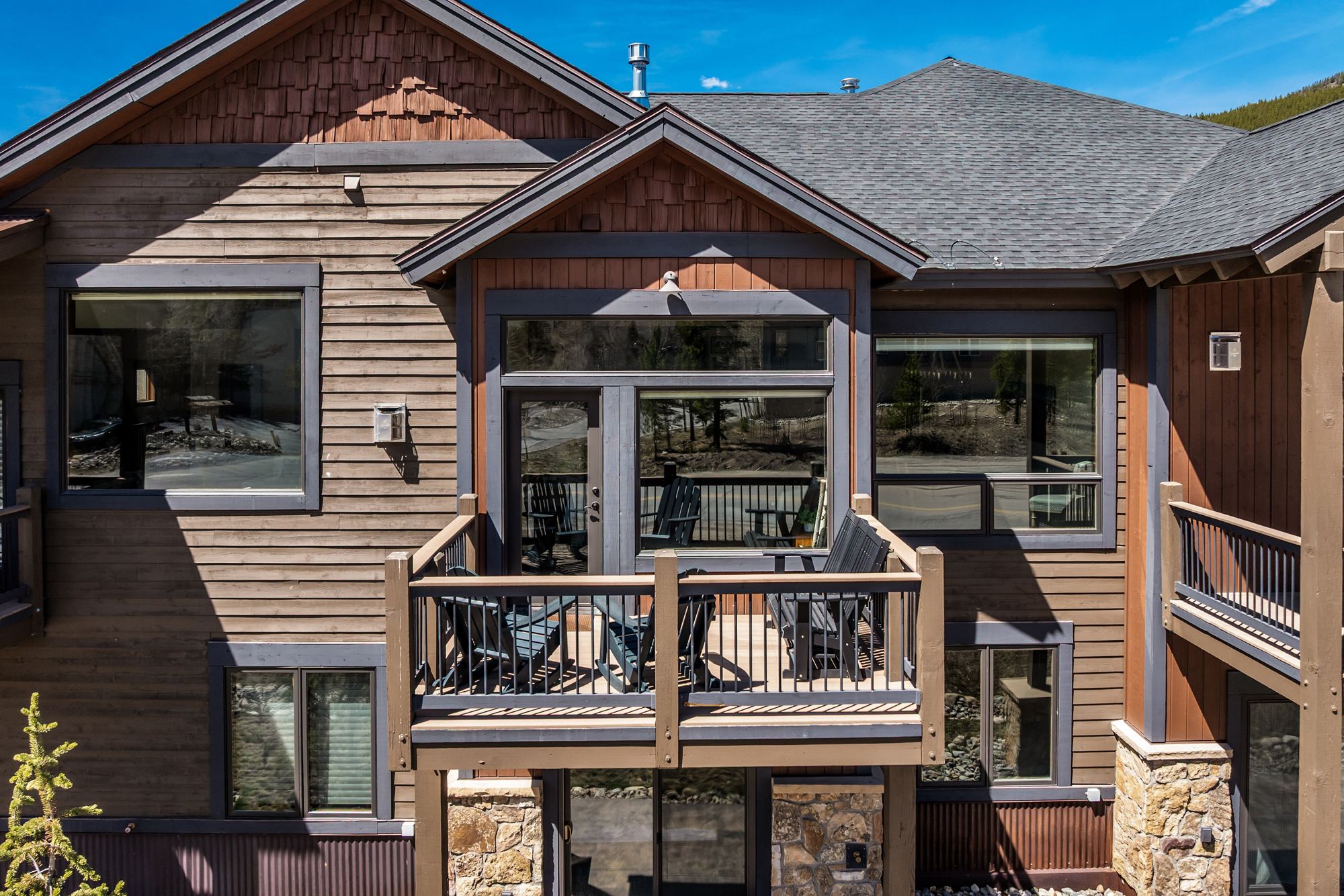 This image captures a charming, elevated wooden balcony attached to a multi-story mountain-style residence. The deck features dark-toned Adirondack chairs and a matching bench, set against a backdrop of horizontal wood siding, stone masonry, and a shingled roof. The perspective is a straight-on, eye-level shot that highlights the architectural details and the inviting outdoor living space.