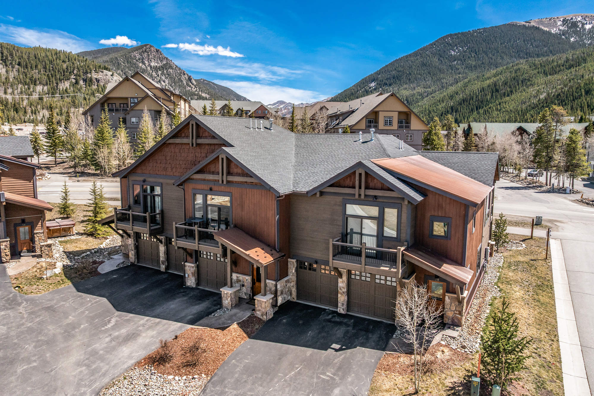 This high-angle aerial shot captures a multi-unit mountain townhome complex featuring a rustic architectural style with wood siding, stone accents, and metal roofing. The property includes multiple garage bays, private balconies, and paved driveways, all set against a stunning backdrop of forested mountains under a clear blue sky. The perspective emphasizes the property's integration into the scenic alpine landscape, highlighting its curb appeal and mountain-resort aesthetic.