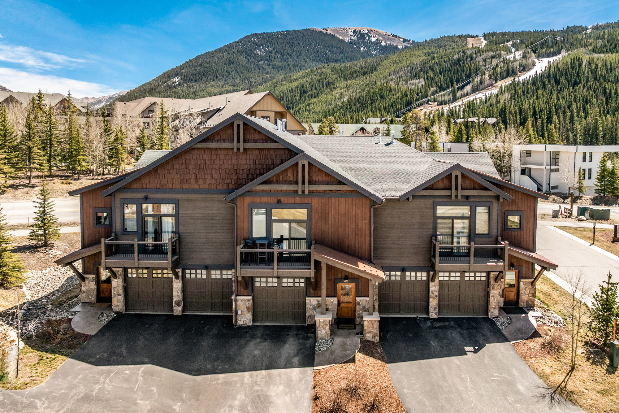 This high-angle exterior shot captures a multi-unit mountain townhome complex featuring a rustic design with wood siding, stone accents, and shingle roofing. The property includes multiple garage bays, private balconies, and paved driveways, all set against a stunning backdrop of a forested mountain range under a clear blue sky. The overall impression is one of a cozy, upscale alpine retreat perfect for mountain living.