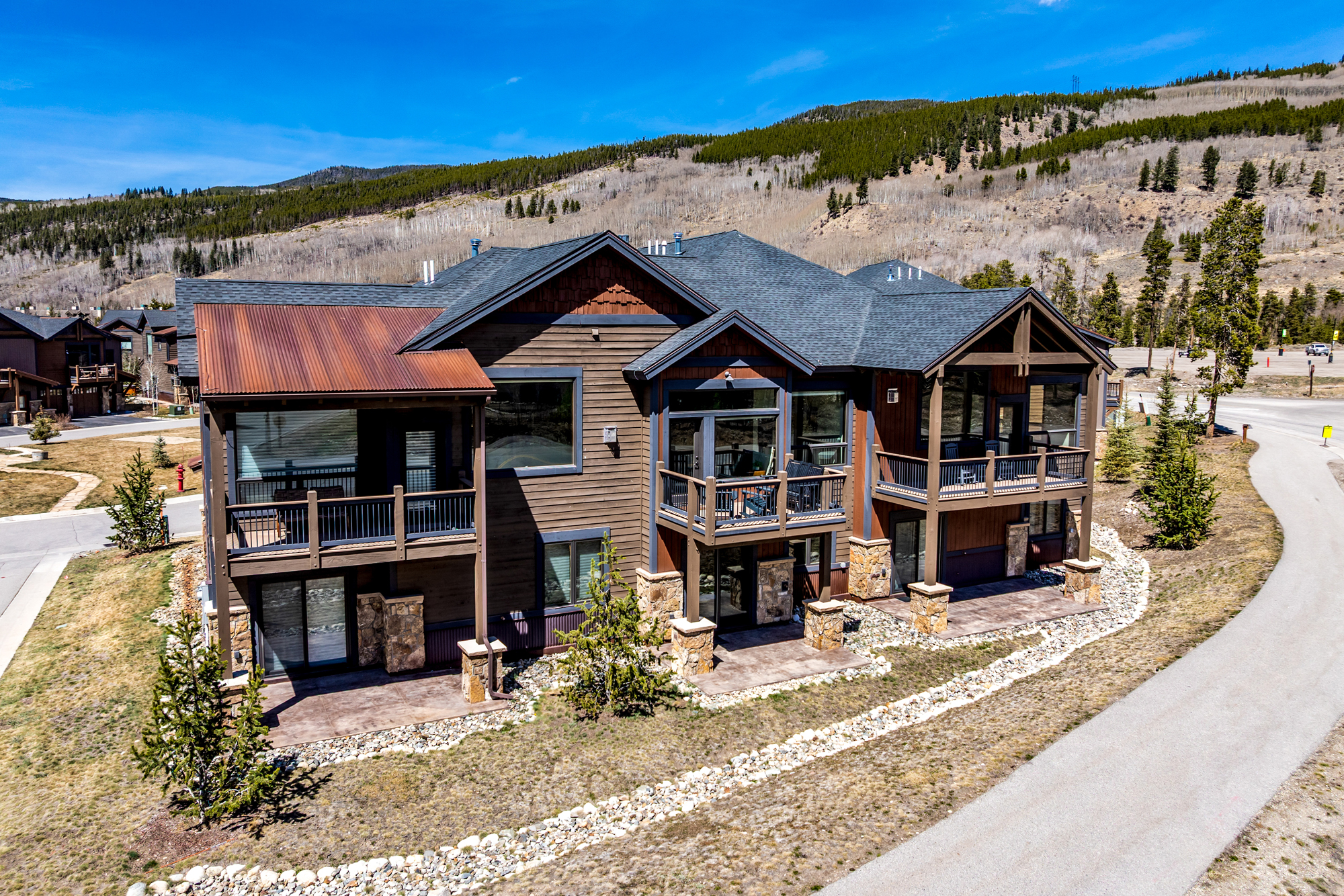 This aerial perspective captures a multi-level mountain-style residence featuring a blend of dark wood siding, stone accents, and a distinctive rusted metal roof section. The home includes multiple balconies and ground-level patios, set against a backdrop of a pine-covered hillside under a clear blue sky. The architecture emphasizes a rustic, lodge-like aesthetic perfectly suited for a high-altitude retreat.