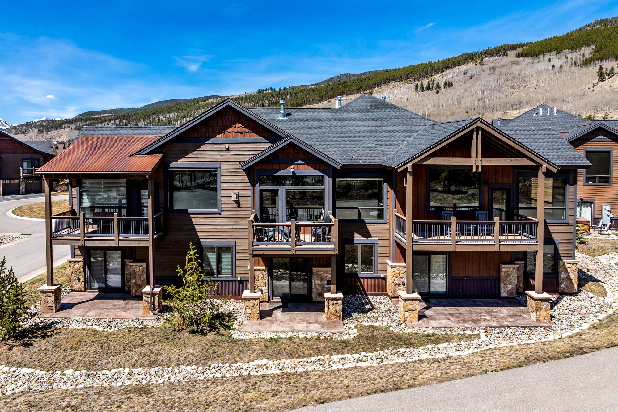 This image showcases the rear exterior of a multi-level mountain-style residence, characterized by its rustic wood siding, stone accents, and a prominent rusted metal roof section. The home features multiple balconies and ground-level patios, offering expansive views of the surrounding landscape and mountain backdrop. The architecture blends seamlessly with the natural environment, creating a warm and inviting mountain retreat aesthetic.