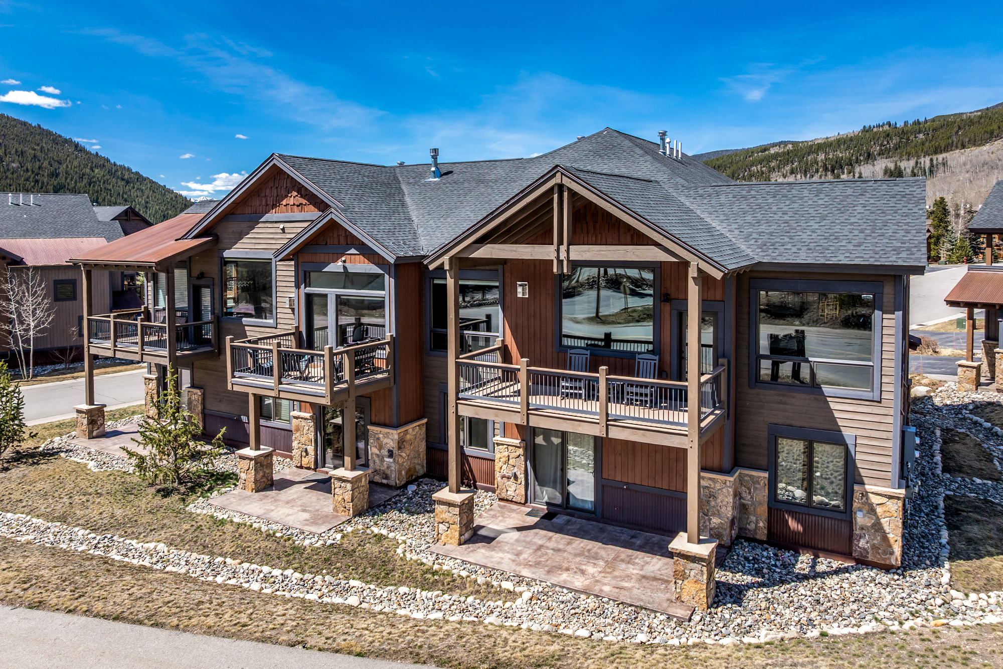 This high-angle exterior shot captures the rear facade of a multi-story mountain-style residence, showcasing a blend of natural stone, wood siding, and shingle roofing. The home features multiple balconies with dark metal railings and ground-level concrete patios, set against a backdrop of clear blue skies and forested hills. The architectural design emphasizes a rustic, lodge-like aesthetic that integrates seamlessly with the surrounding rocky landscape.
