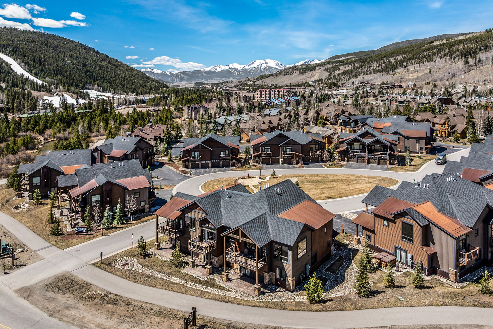 This high-angle aerial view captures a picturesque mountain residential community featuring multi-story townhomes with rustic wood siding and dark shingled roofs. The neighborhood is nestled in a valley surrounded by lush pine forests and snow-capped peaks, creating a serene and upscale alpine atmosphere. A paved road winds through the development, highlighting the well-maintained landscaping and proximity to nature.