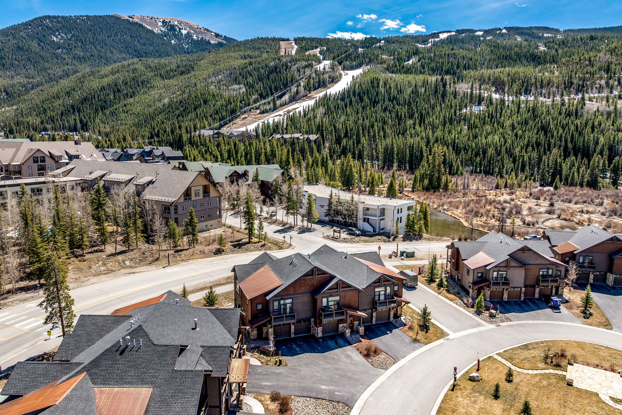 This high-angle aerial shot captures a scenic mountain residential community featuring multi-story townhomes with dark shingled roofs and rustic wood siding. The perspective highlights the neighborhood's layout, including paved driveways and roads, set against a backdrop of a lush, forested mountain slope with a visible ski run. The overall impression is one of a peaceful, upscale mountain retreat perfect for outdoor enthusiasts.