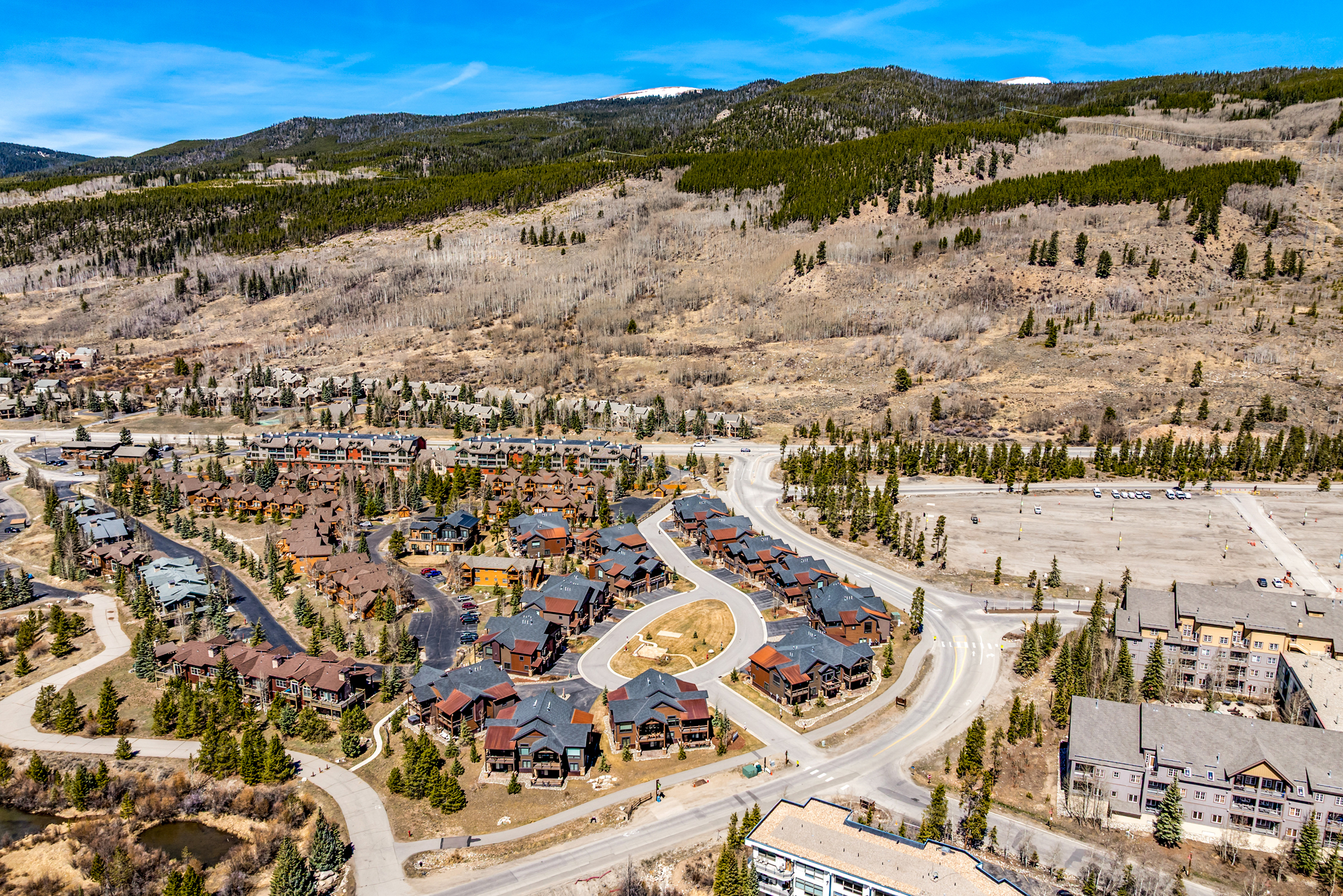 This high-angle aerial view captures a sprawling mountain resort community nestled at the base of a vast, forested hillside. The scene features numerous multi-story residential buildings with dark shingled roofs, interconnected by winding paved roads and surrounded by evergreen trees and dry, seasonal vegetation. The perspective provides a comprehensive look at the neighborhood's layout, emphasizing its serene, secluded location and proximity to nature.