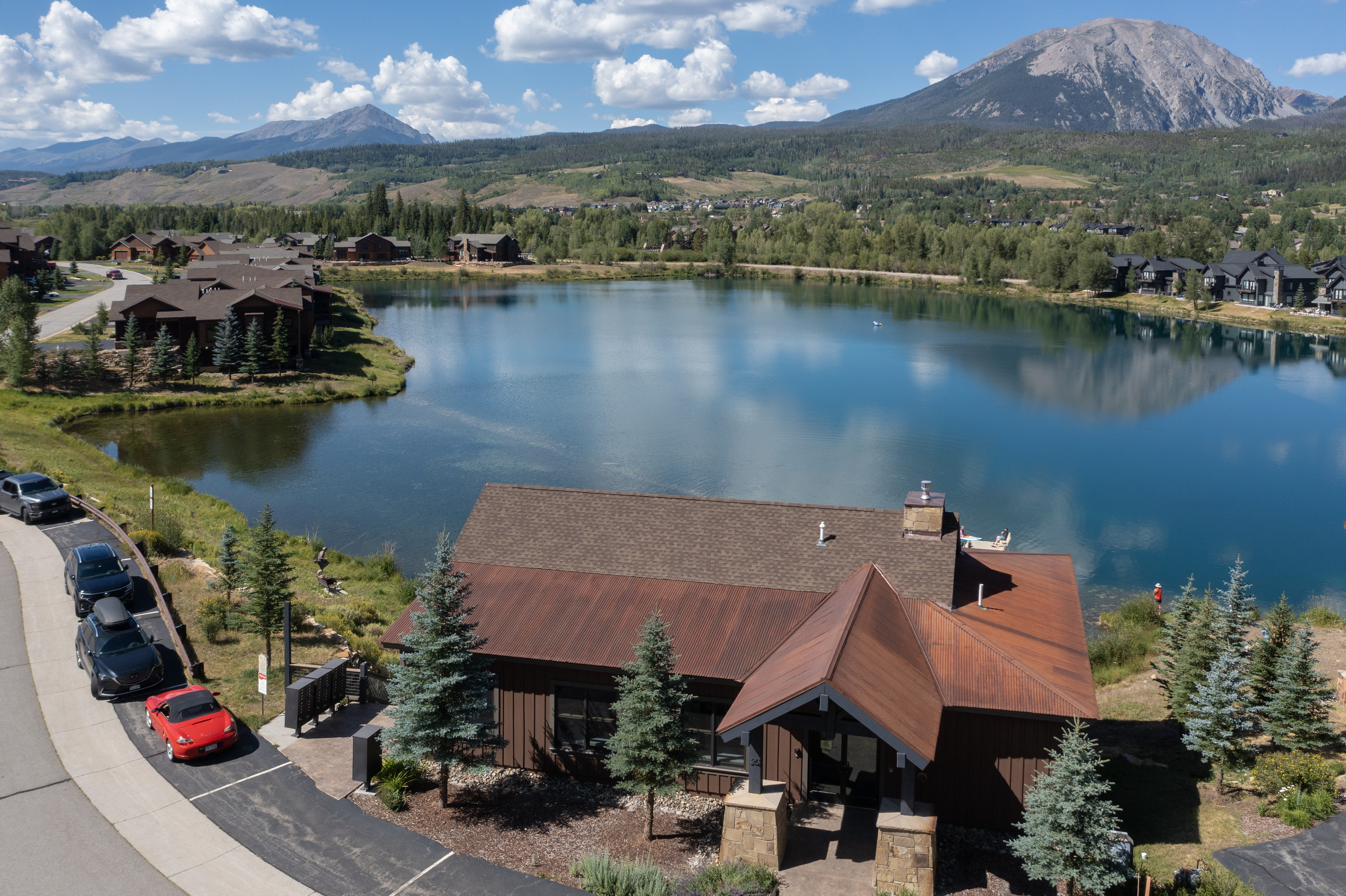 This high-angle aerial shot captures a mountain-style building situated directly on the edge of a serene, deep blue lake. The structure features a distinctive rust-colored metal roof, dark wood siding, and stone accents, complemented by a row of parked vehicles along the adjacent paved road. In the background, majestic mountain peaks rise against a clear sky, creating a picturesque and tranquil setting for this waterfront property.