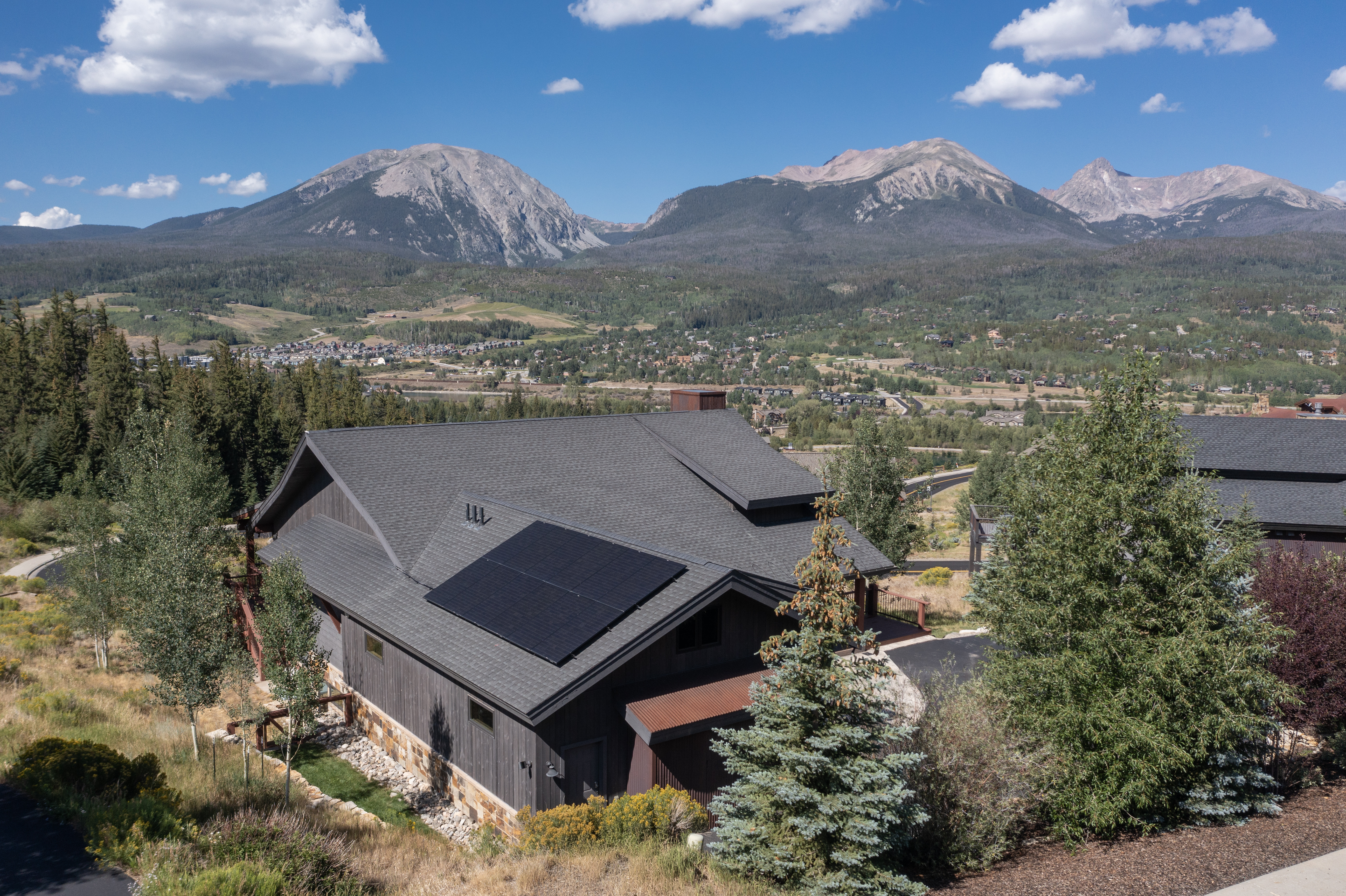 This high-angle aerial shot captures a modern mountain home nestled in a scenic valley with dramatic, snow-dusted peaks in the background. The residence features dark vertical siding, a multi-pitched roof with integrated solar panels, and a stone foundation, blending seamlessly with the surrounding evergreen forest. The expansive landscape provides a serene, elevated perspective of the property's integration into its rugged, natural environment.
