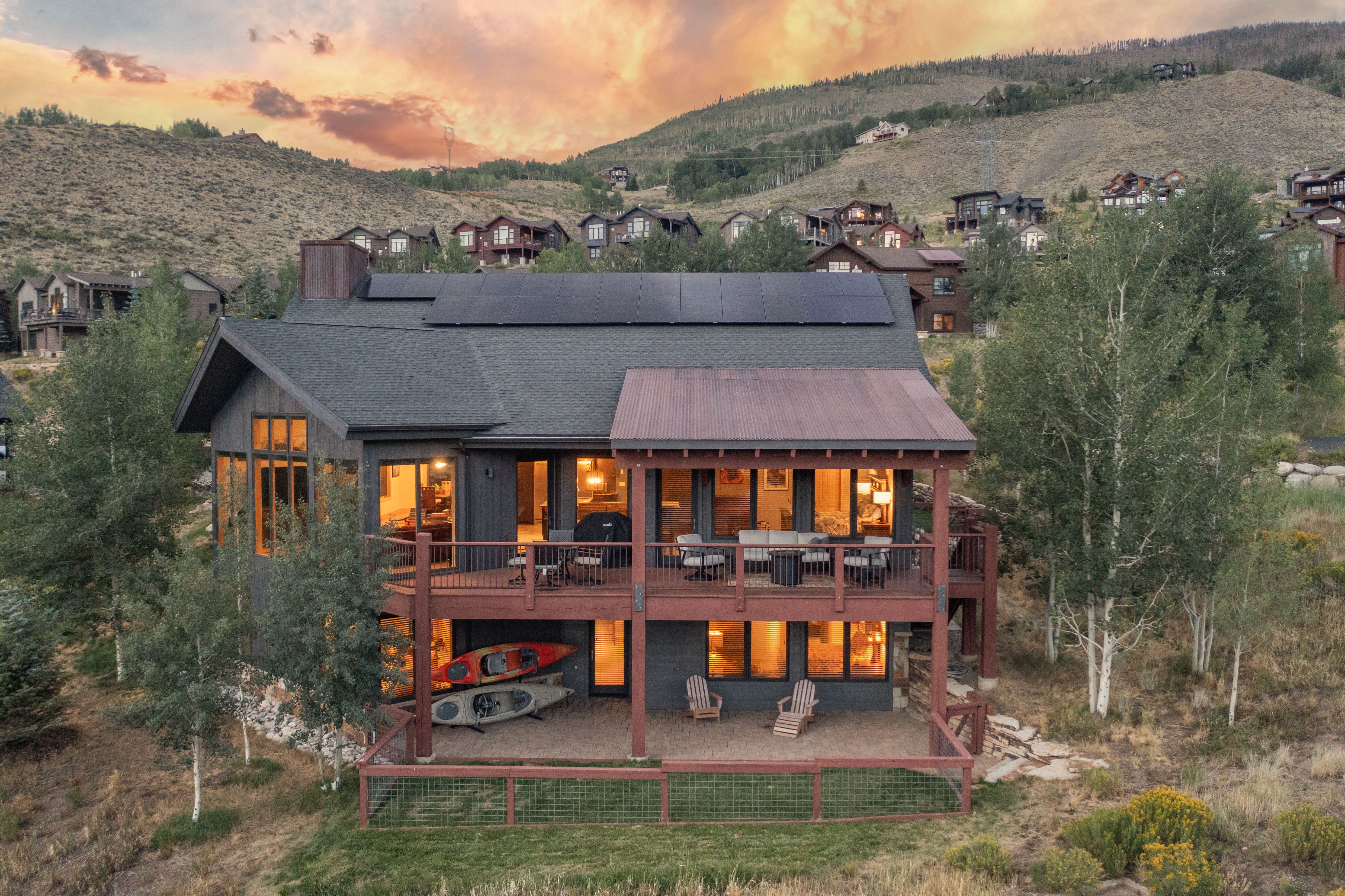 This elevated rear view captures a multi-level mountain home set against a dramatic sunset sky. The residence features dark wood siding, a large upper deck with outdoor seating, and a lower covered patio area equipped with two chairs and stored kayaks. Solar panels are installed on the roof, and the property is surrounded by a natural landscape of trees and rolling hills.